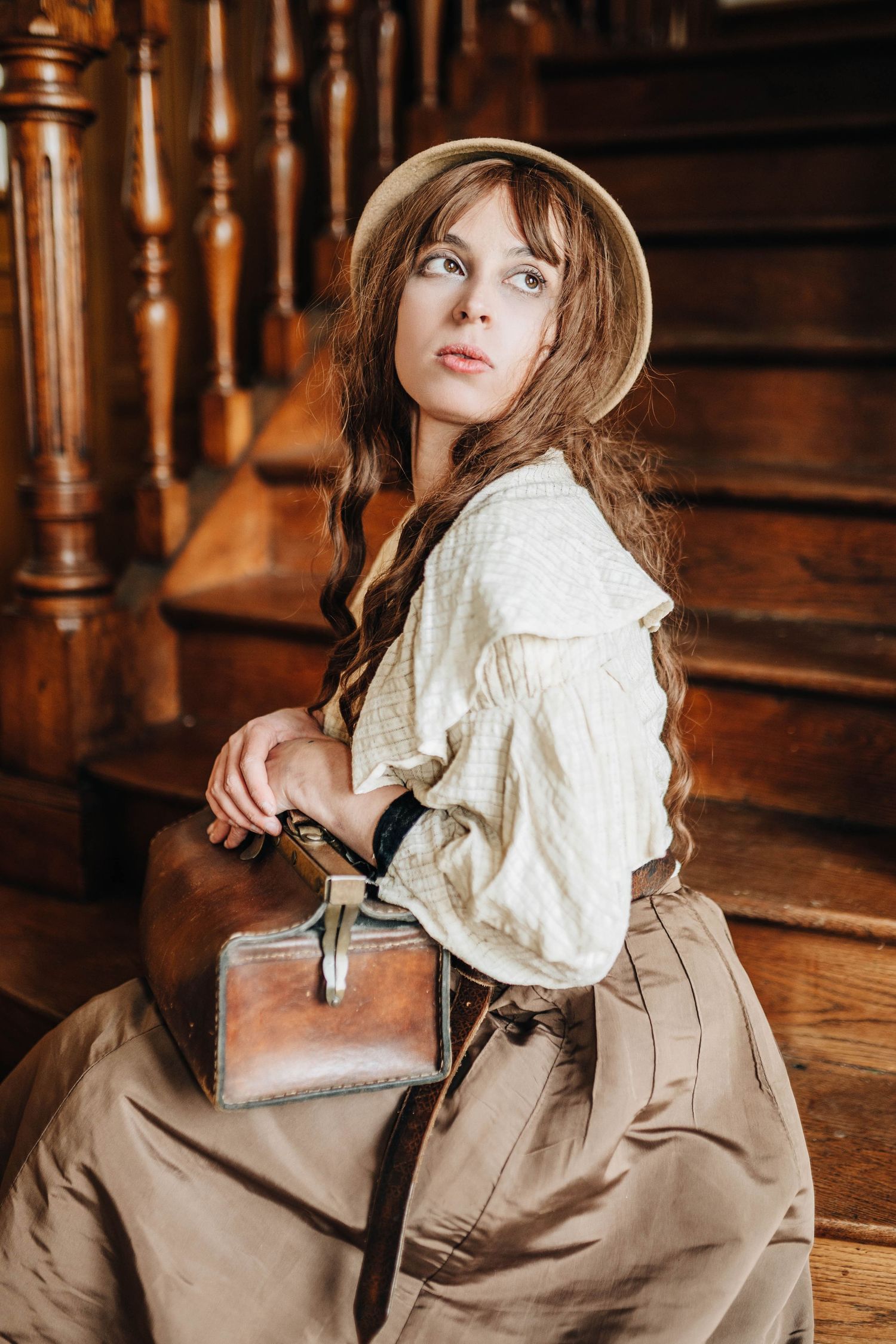 Young woman in vintage attire with hat and leather bag sits on wooden stairs in a period-style portrait.