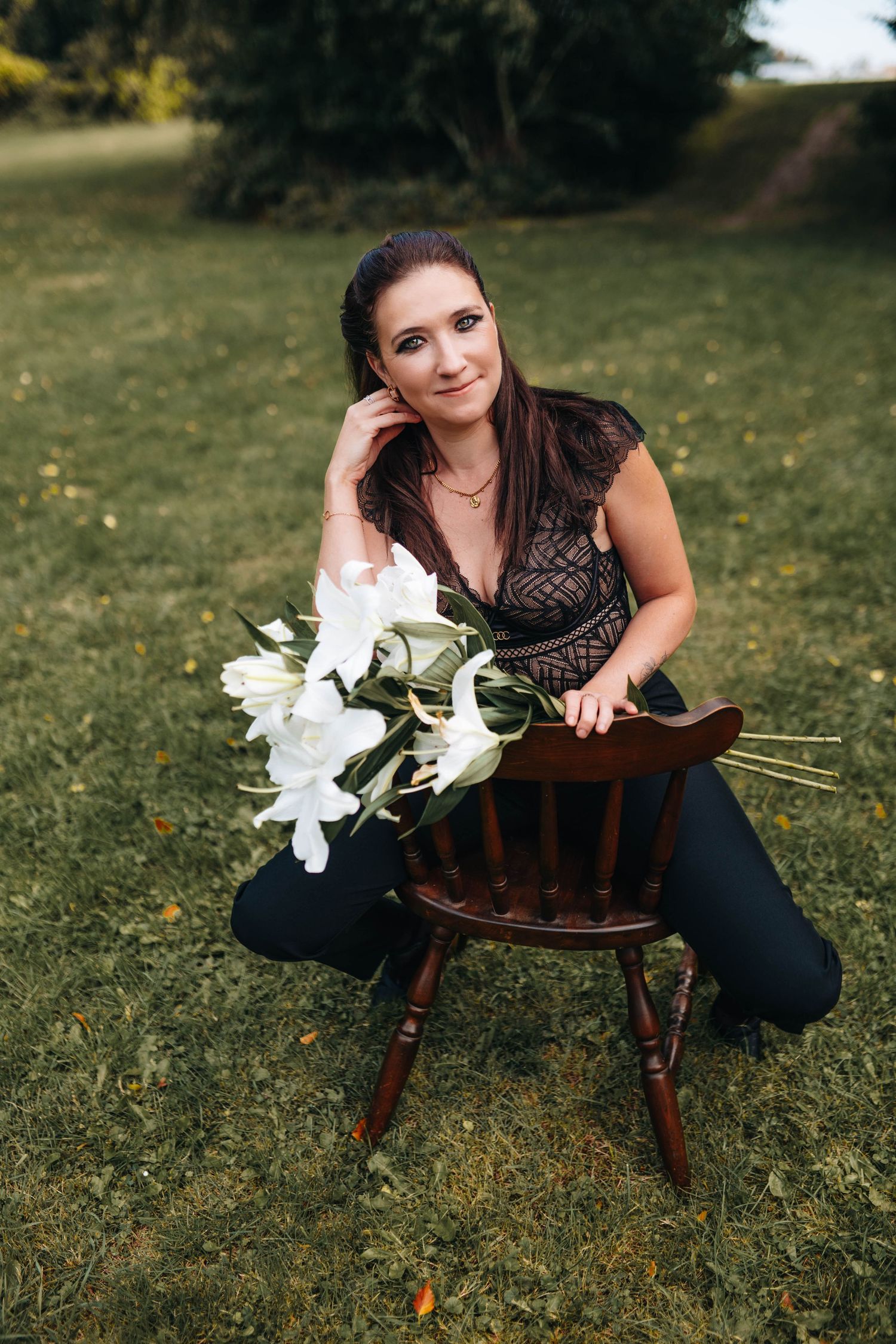 Woman in black lace top poses with white lilies on wooden chair in garden setting.