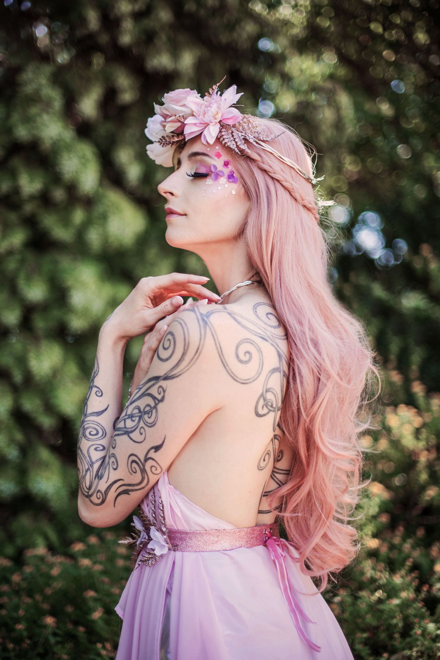 A person with artistic tattoos poses in a pink dress among garden foliage.