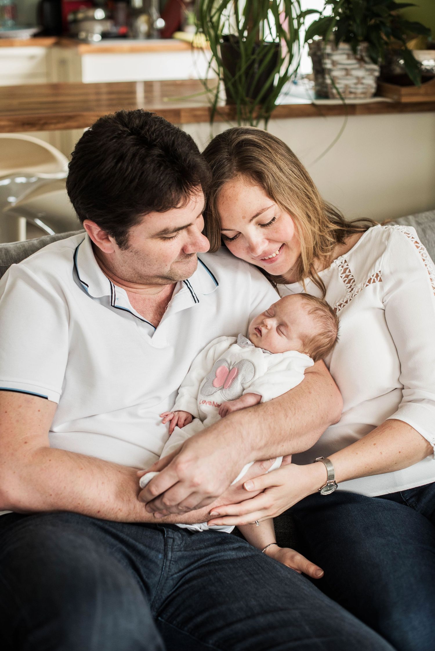 Parents in white shirts cuddling with newborn on couch.