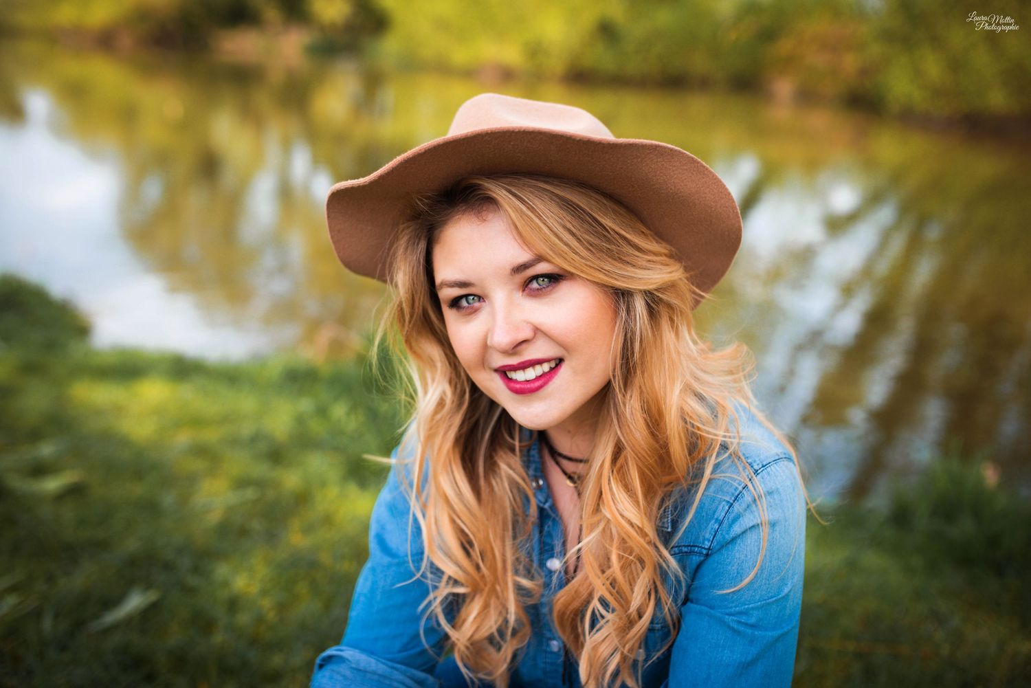 Outdoor portrait by a pond wearing denim shirt and brown hat in natural lighting.