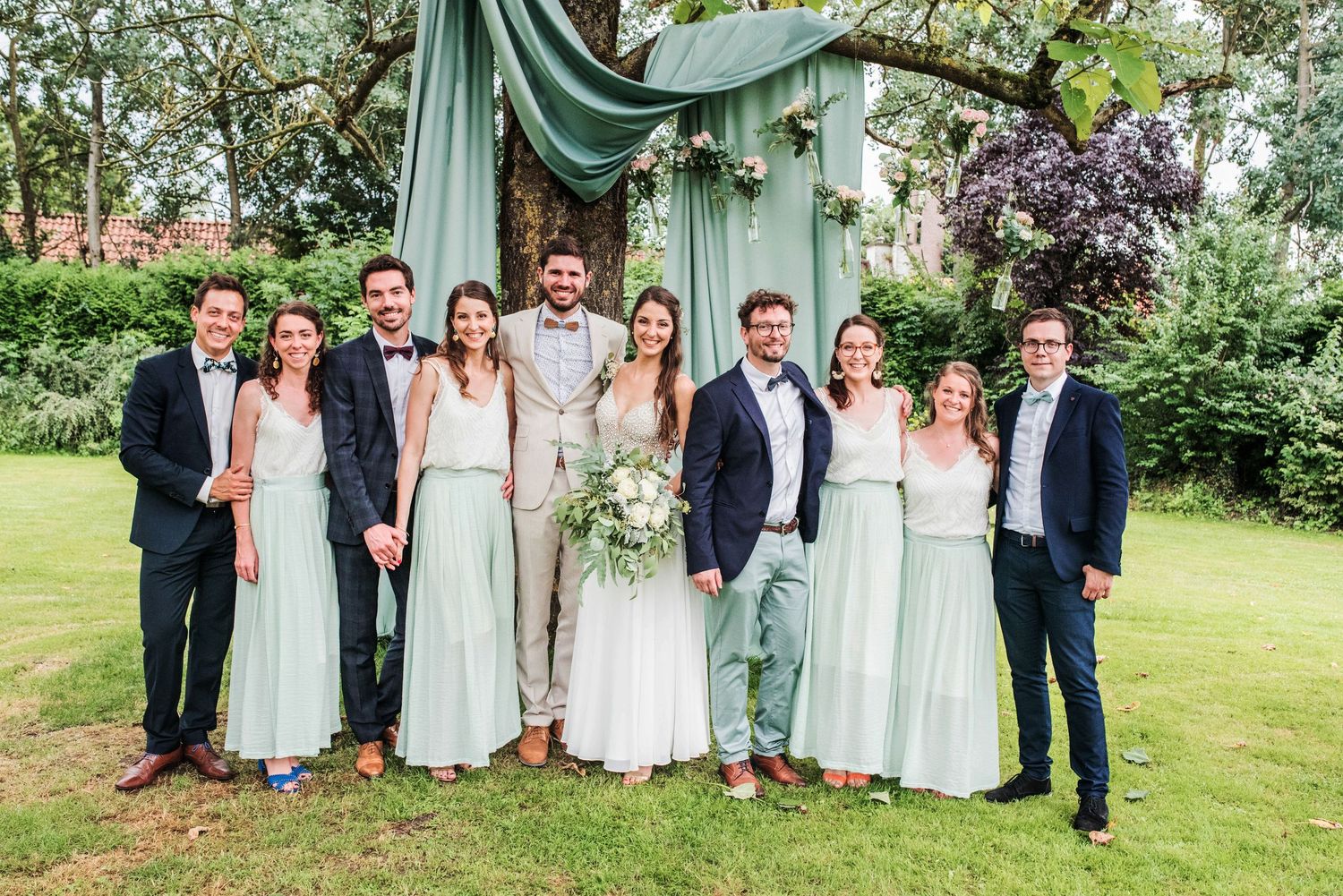 Wedding party in mint green and gray attire poses together on a lush garden lawn.