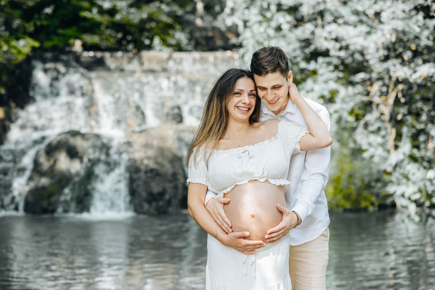 Pregnant couple embracing outdoors near waterfall with blooming white trees, woman in white dress cradling baby bump.