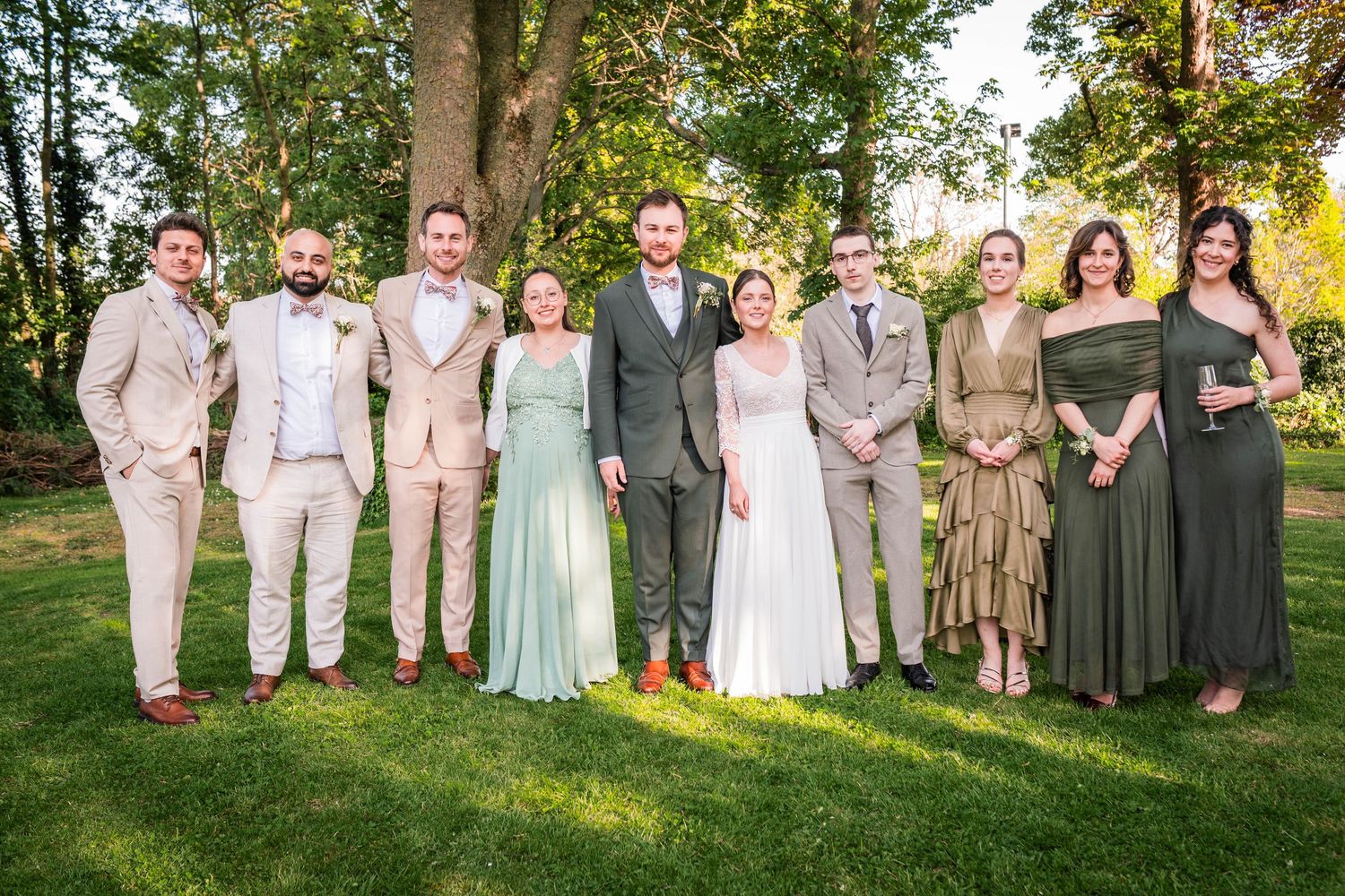 Wedding party posing outdoors on green lawn, bride in white, groom in gray, bridesmaids in green and gold dresses.