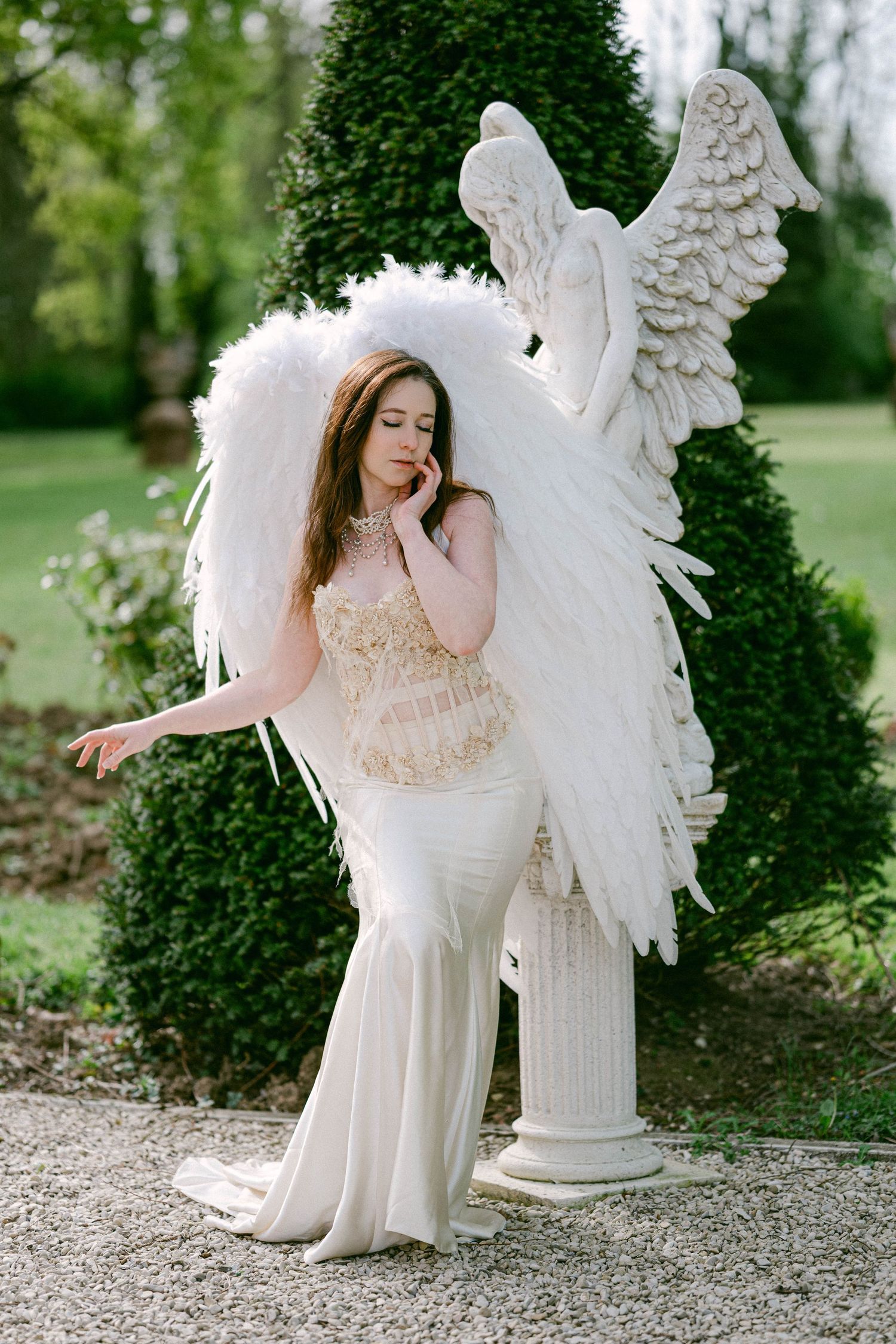 Woman in white angel costume with large feather wings poses beside an angel statue in a garden.