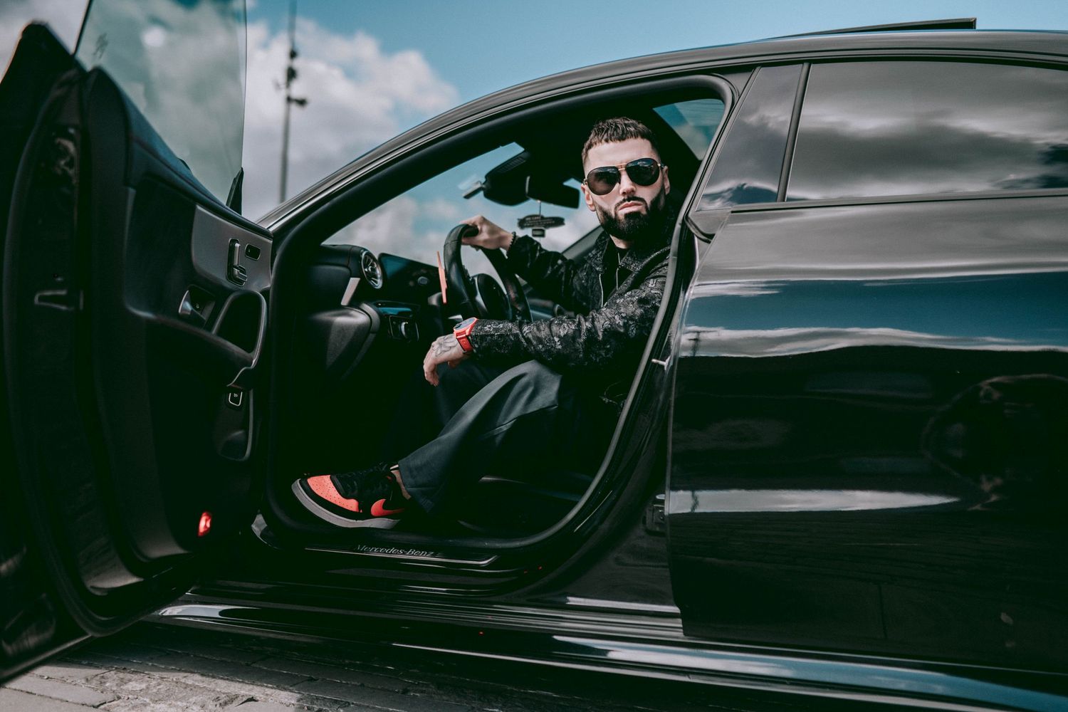 A stylish man wearing sunglasses sits in the driver seat of a sleek black luxury car with the door open.