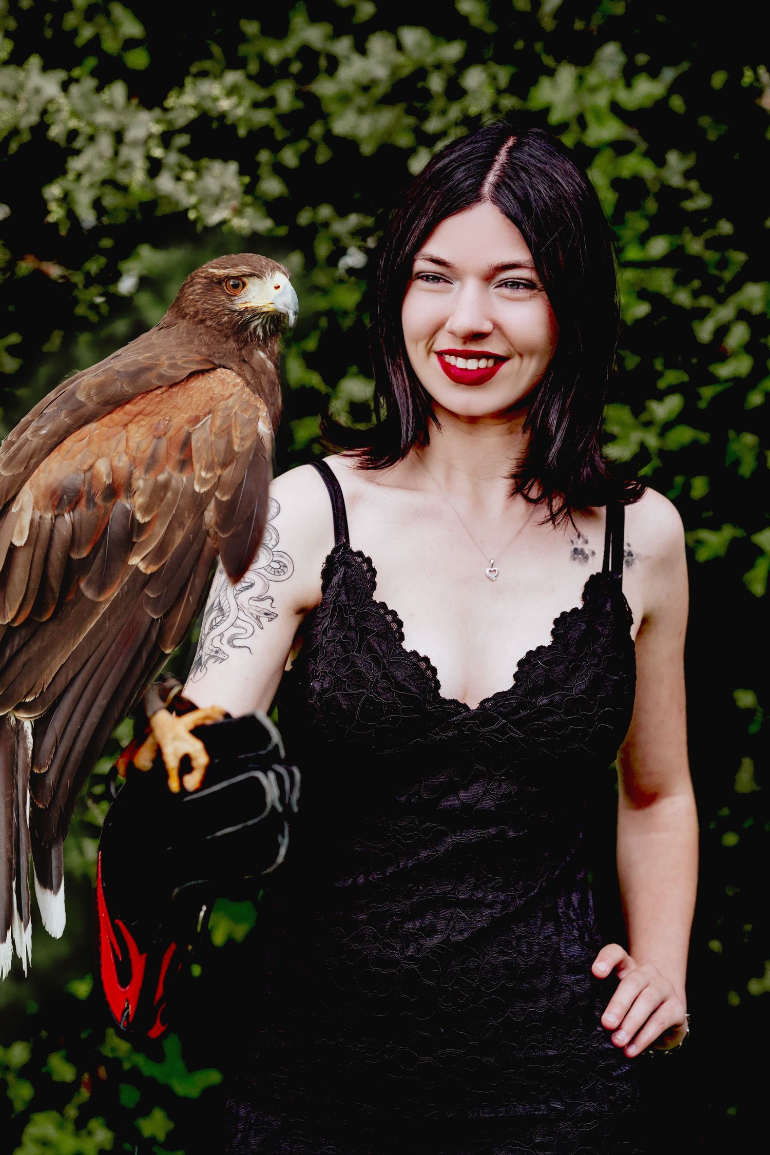 Woman in black lace dress with red lips holds a hawk on her gloved arm, smiling outdoors near green foliage.