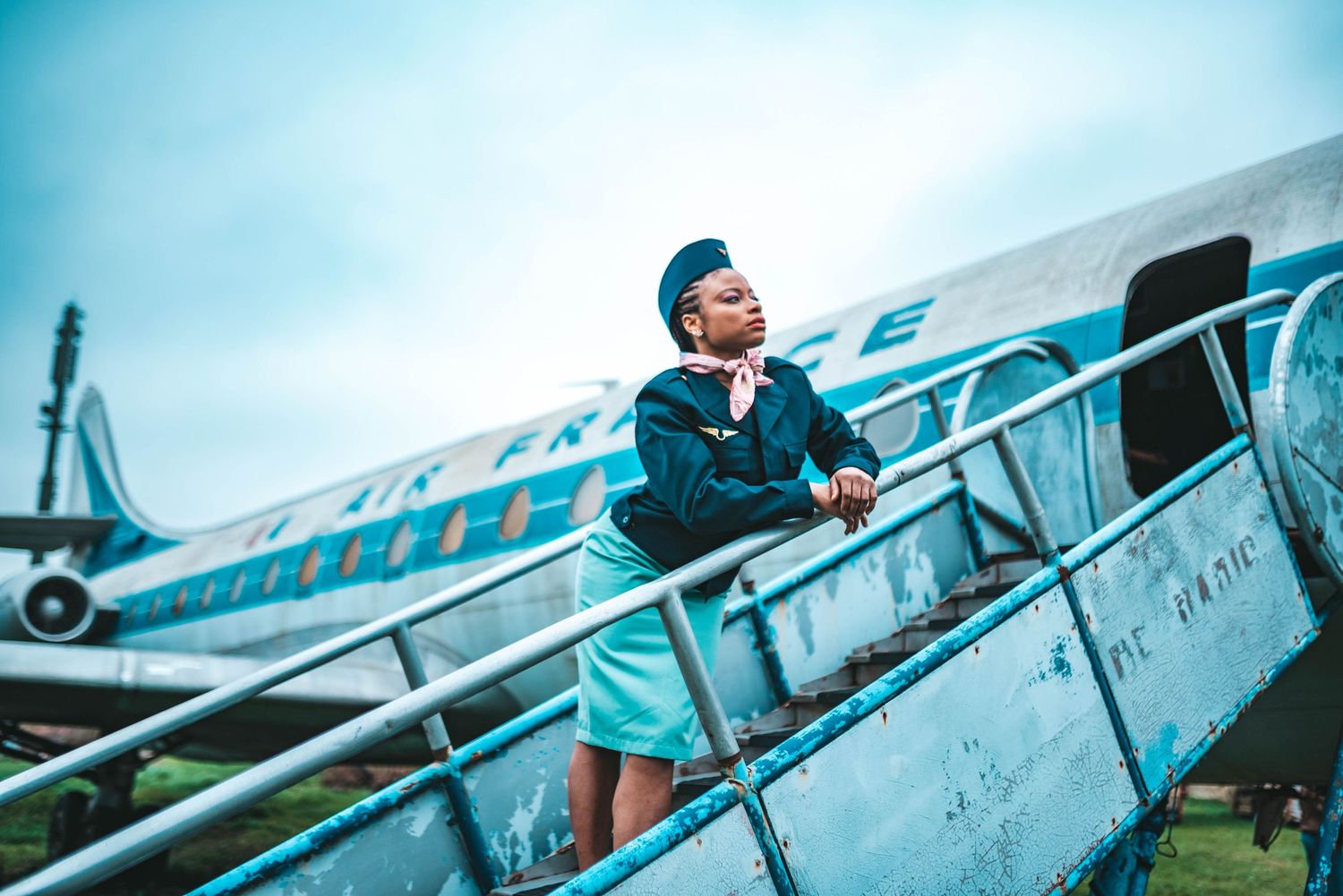 Flight attendant in teal uniform poses on airplane boarding stairs against blue sky.