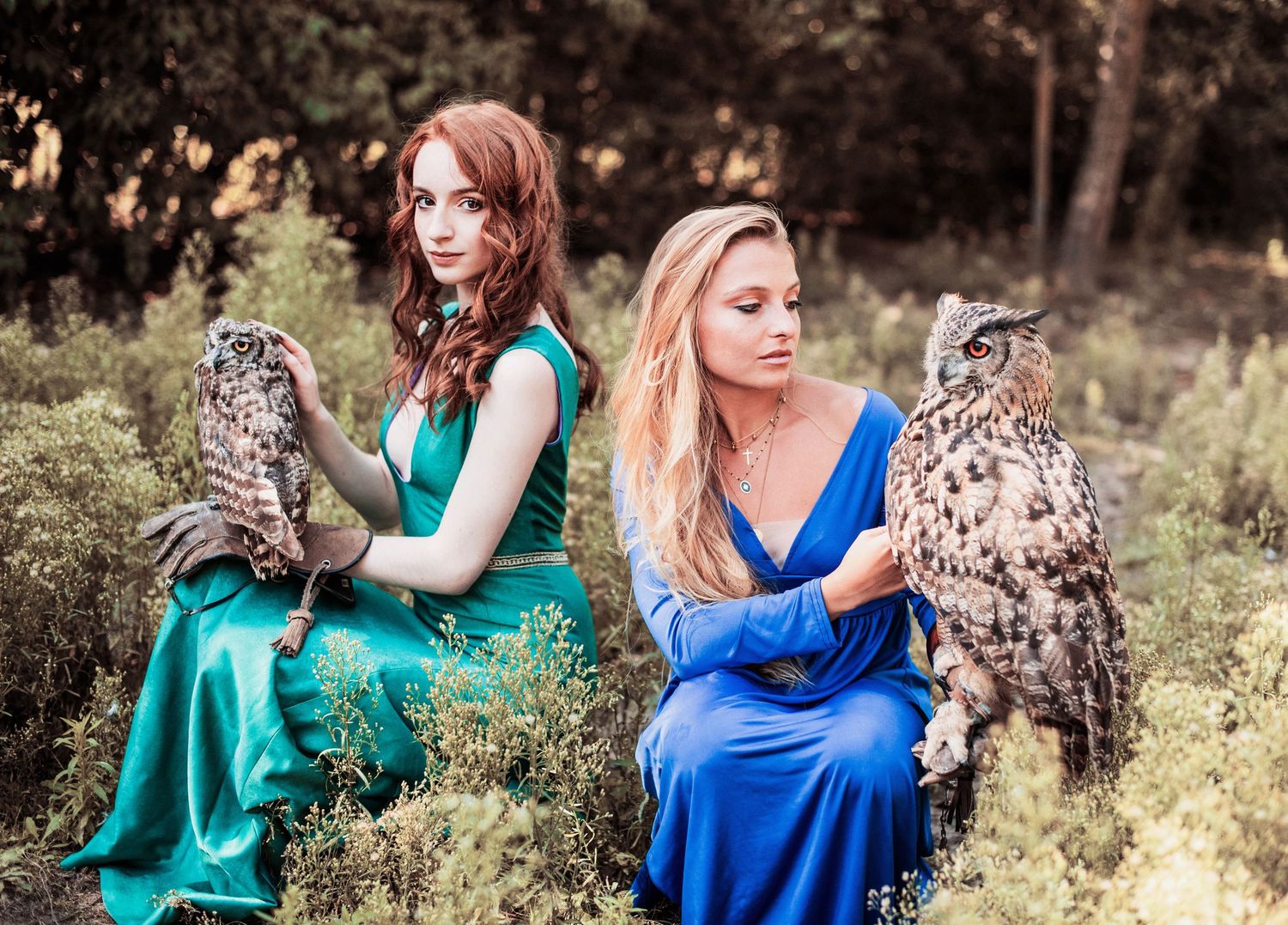 Two models in flowing gowns pose with owls in a magical forest photoshoot.