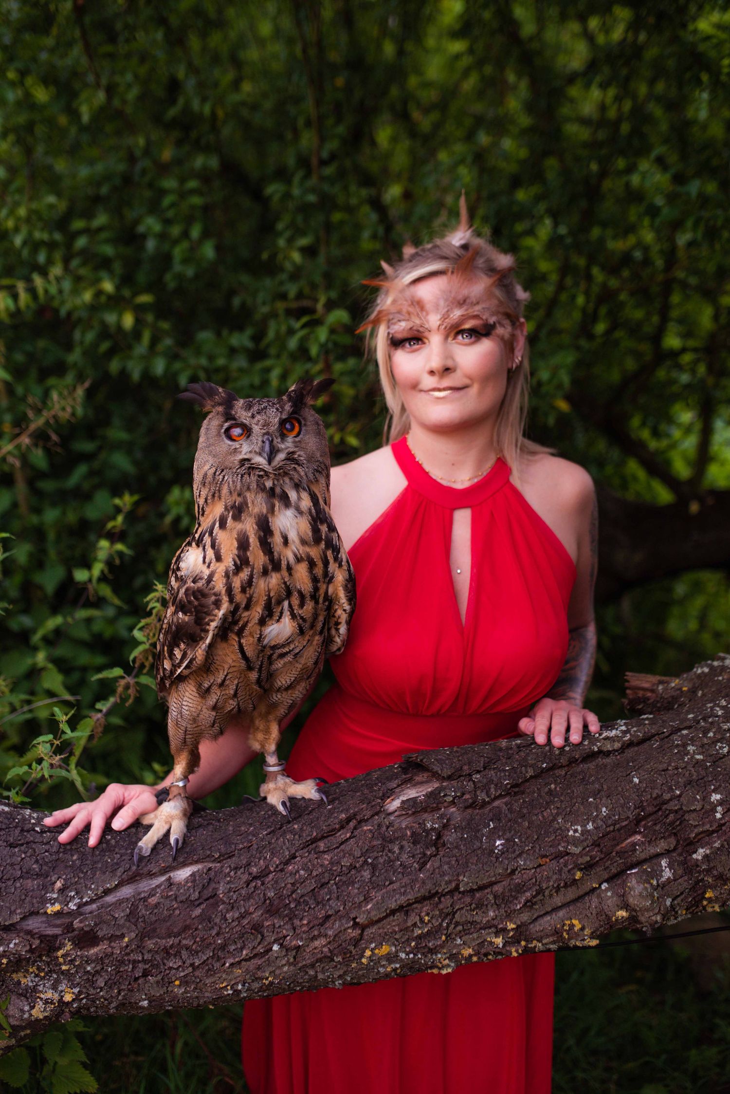 A large owl and its handler pose together on a fallen log in a forest setting.