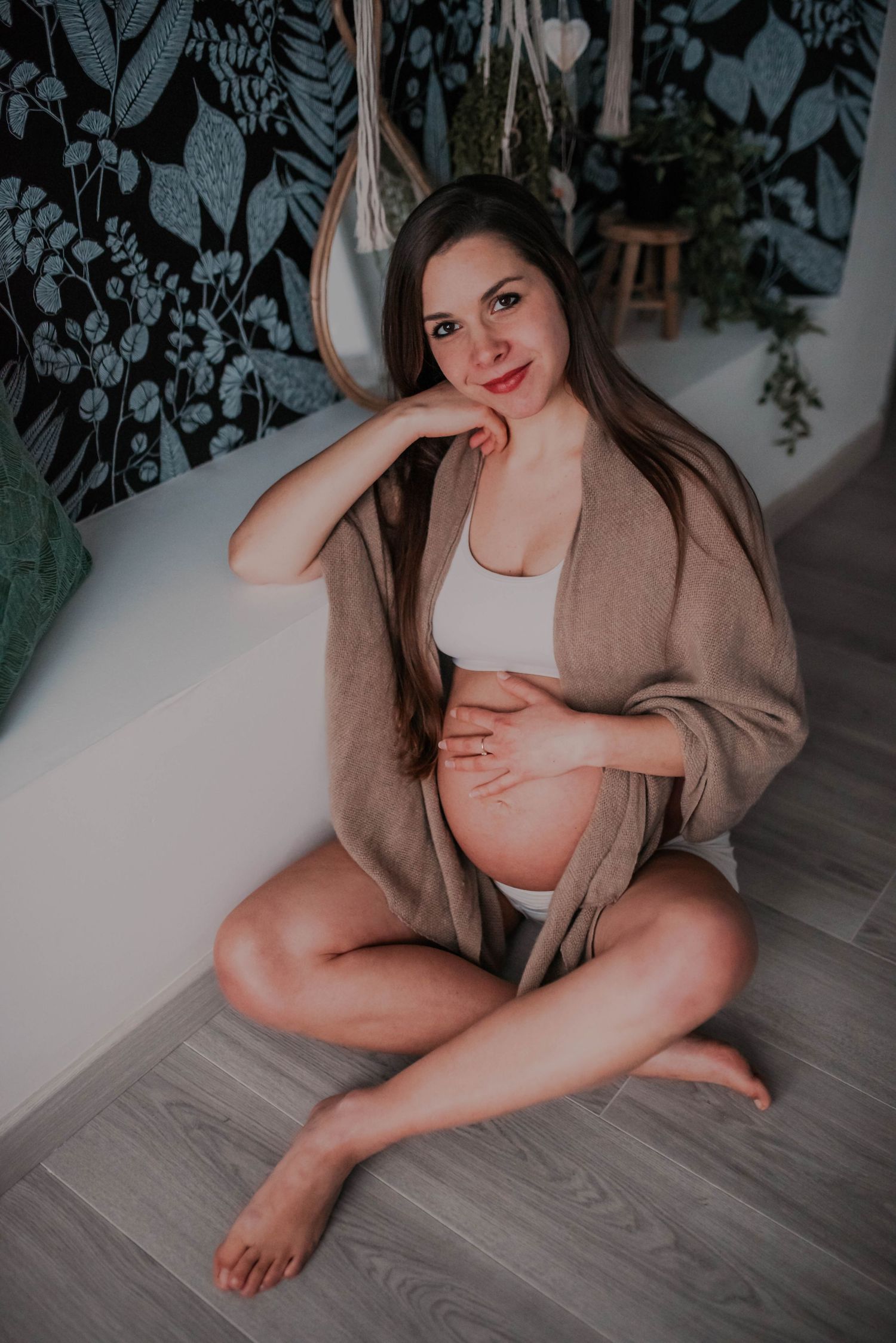 Indoor maternity boudoir session with cozy cardigan and floral wallpaper backdrop.