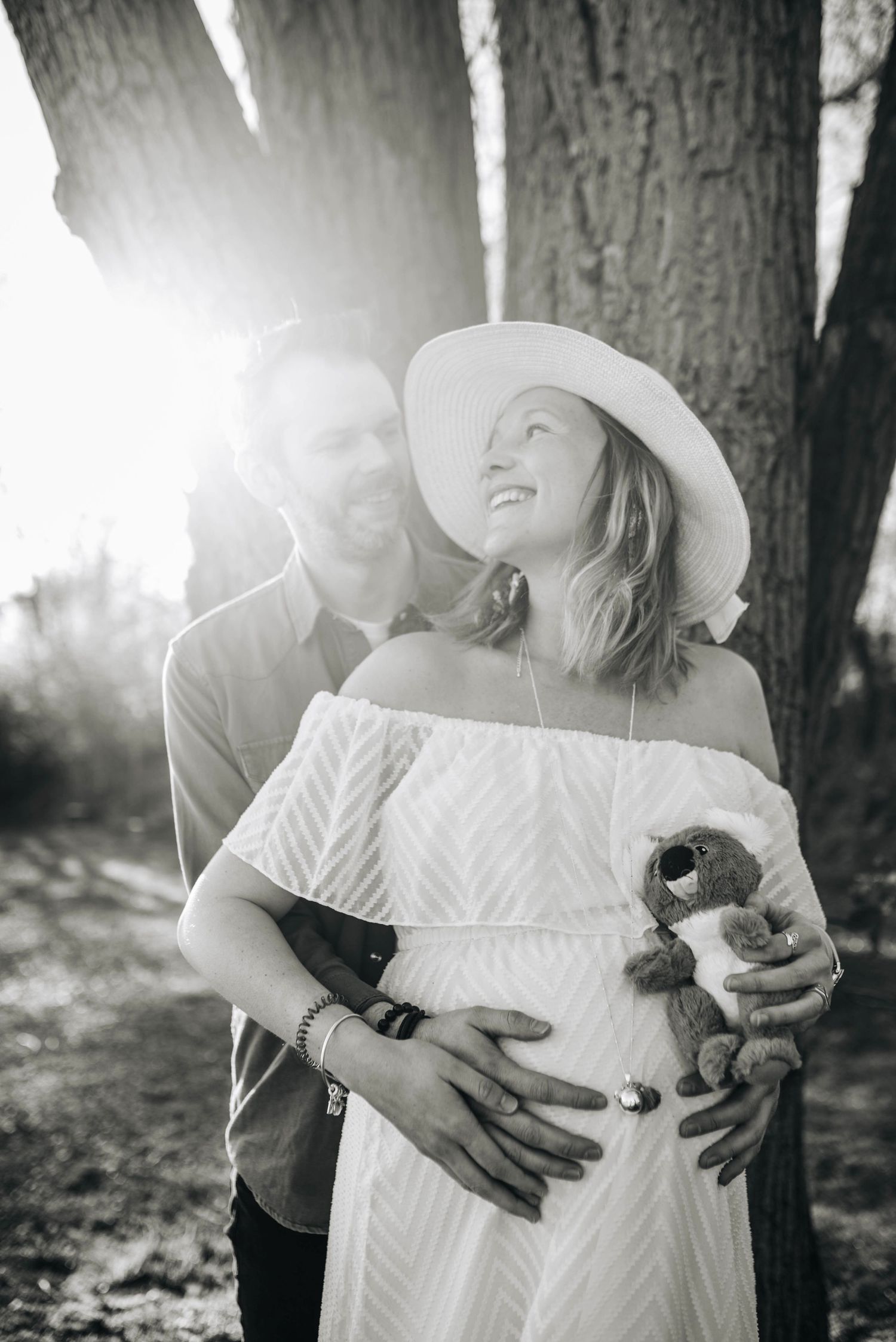 A sunlit maternity moment captured under large trees with a white summer dress.