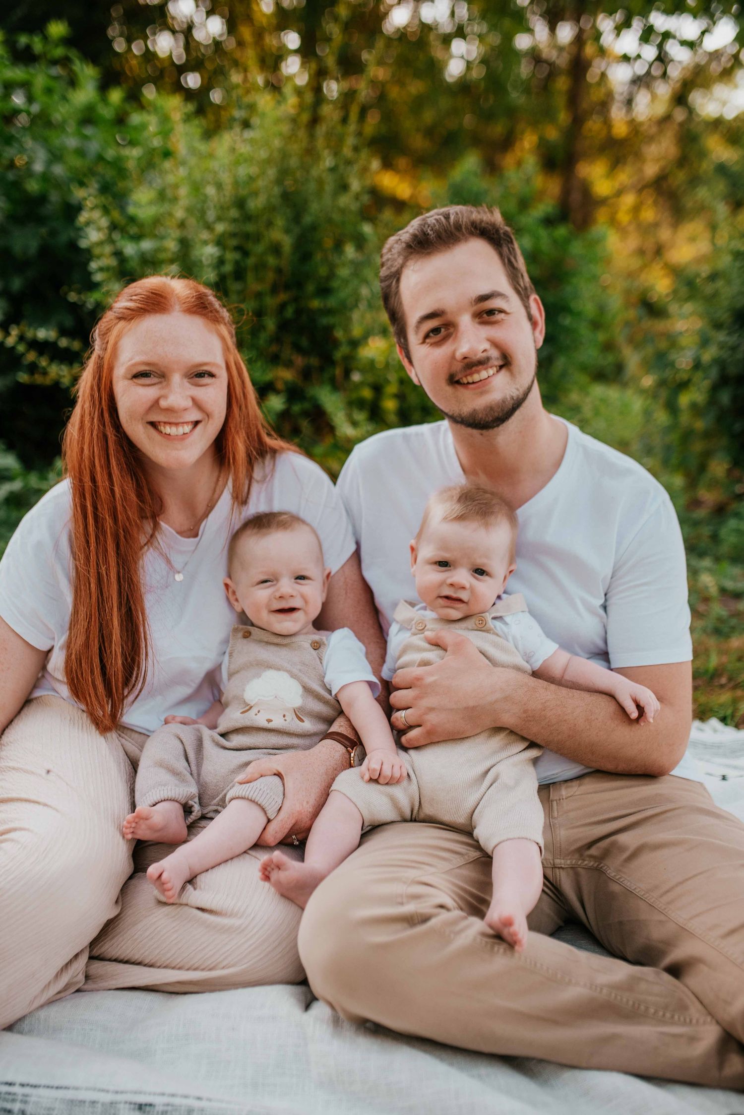 Family relaxing together on white blanket outdoors in neutral colored clothing.