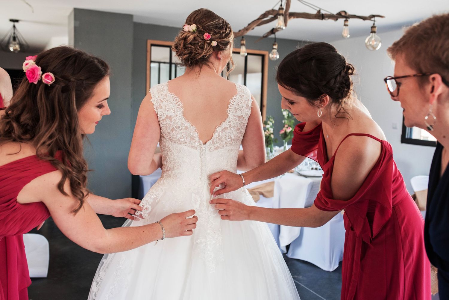 Bridesmaids in burgundy dresses help with wedding preparations in a getting ready room.