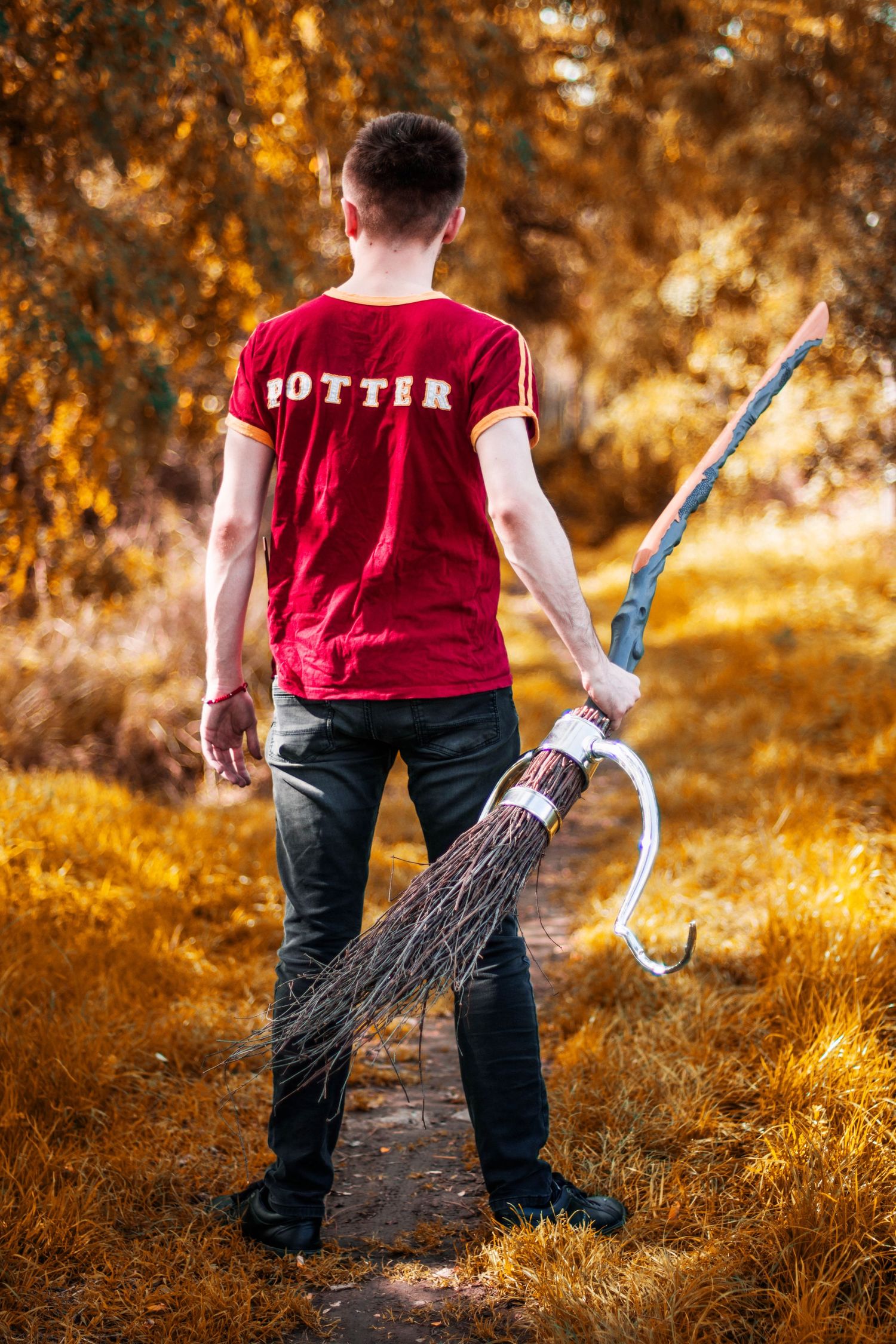 Someone wearing a red Potter shirt poses with a broomstick in a golden autumn setting.