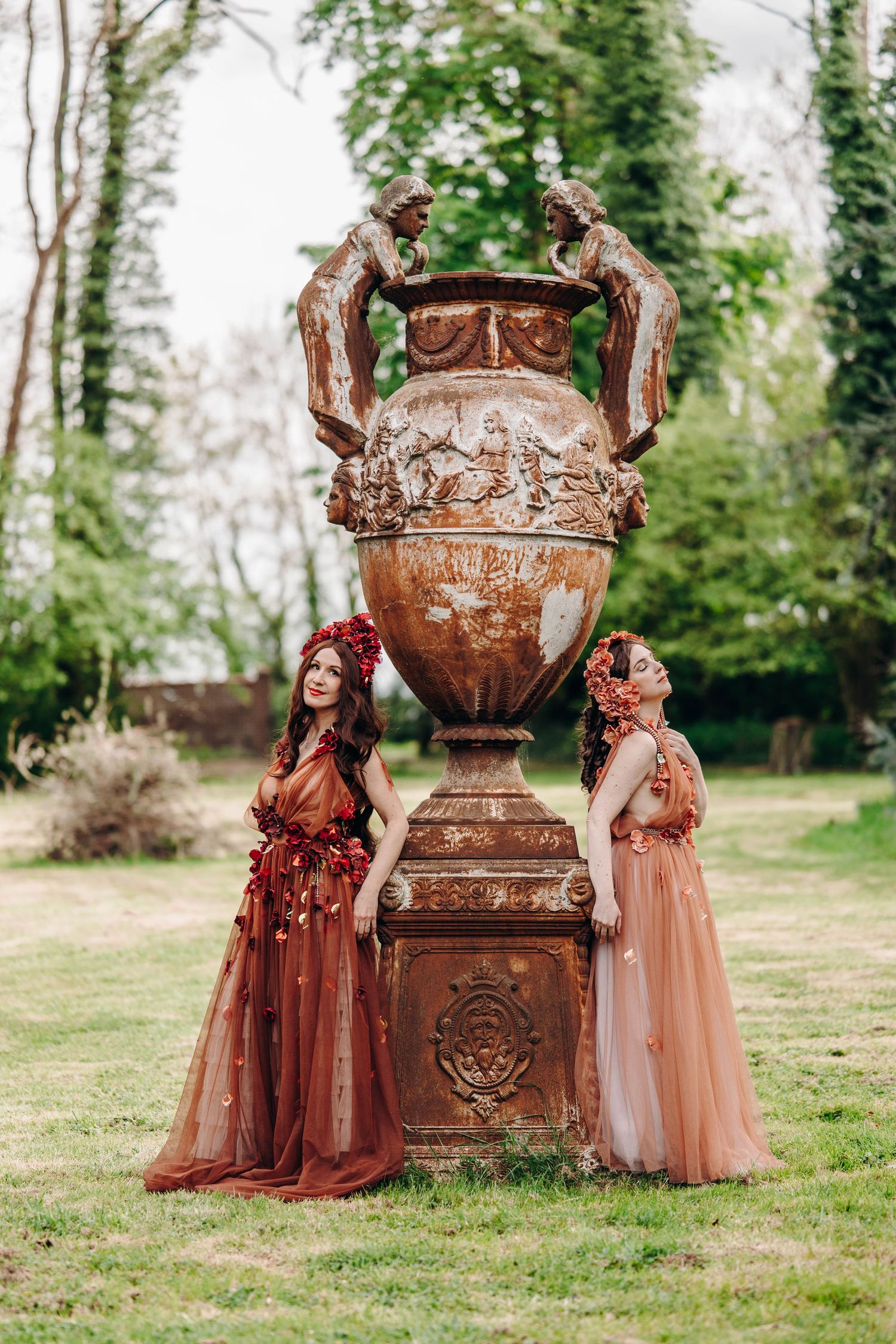 Two women in elegant rust-colored gowns and floral crowns pose beside a large ornate antique urn in a lush garden.