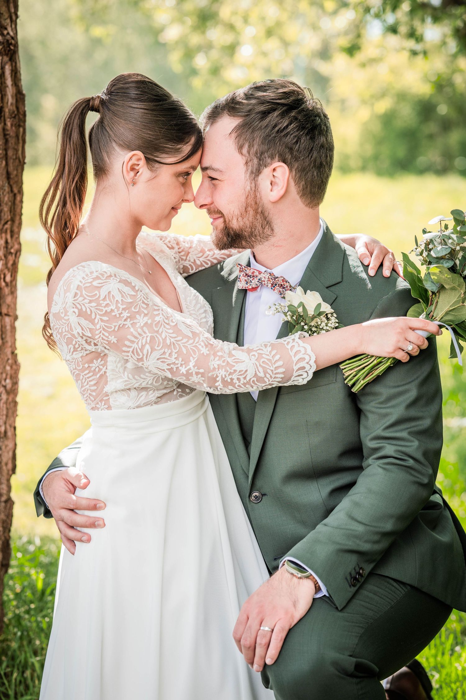 A bride and groom share a romantic moment outdoors, with the groom in a green suit and bride in a lace wedding dress.