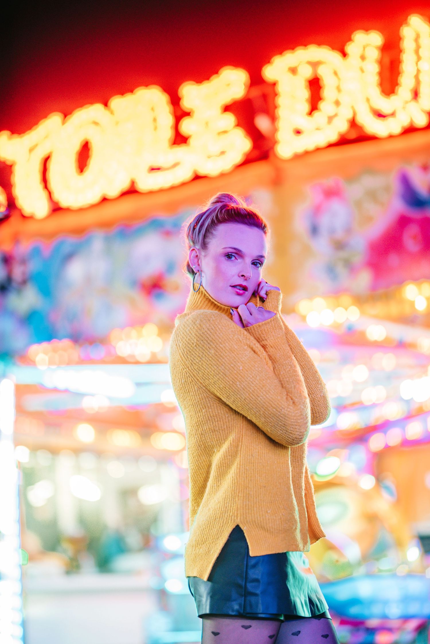 Fashion portrait with bright neon carnival lights and yellow sweater creating vibrant nighttime ambiance.