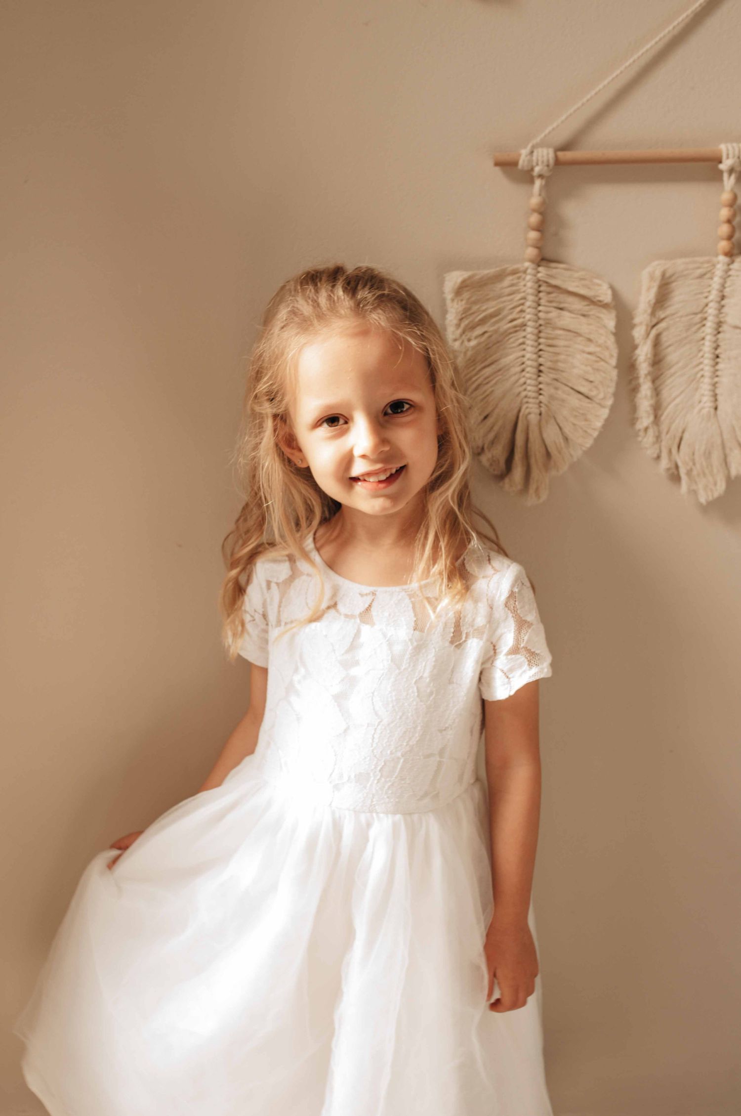Young girl in white lace dress posing against neutral wall with macrame decor.