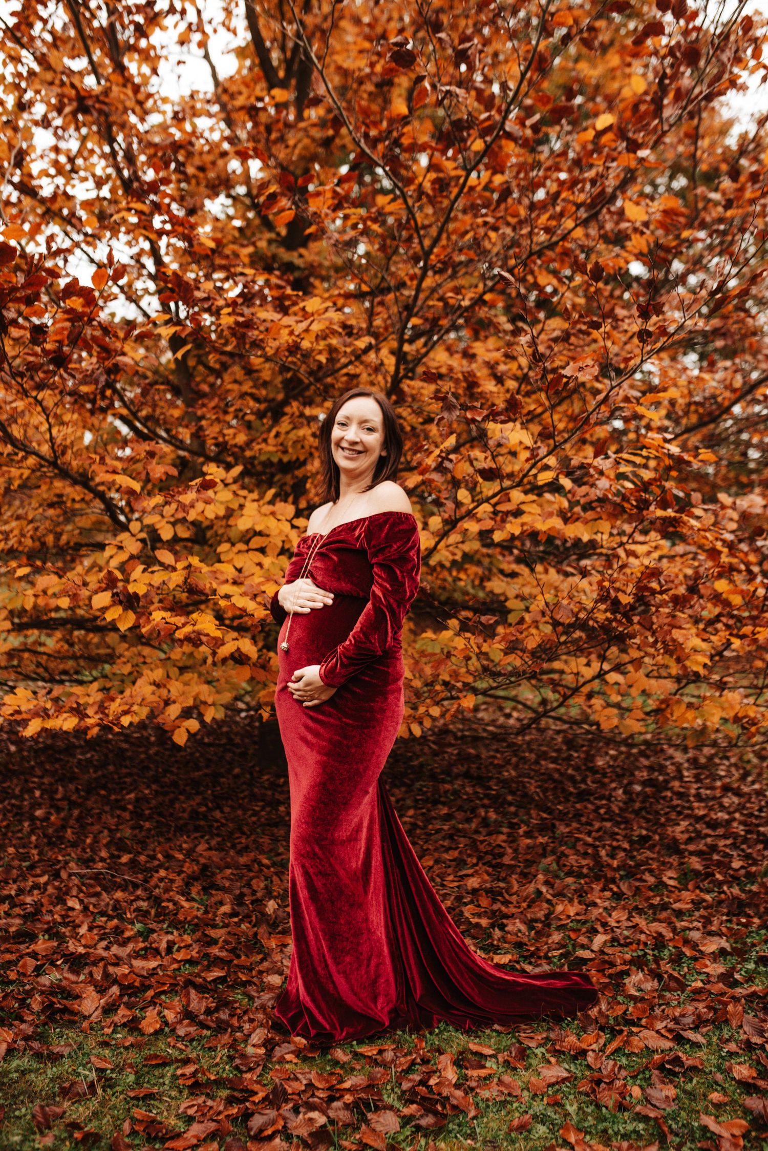 A figure in a flowing burgundy velvet dress poses against vibrant orange autumn foliage.