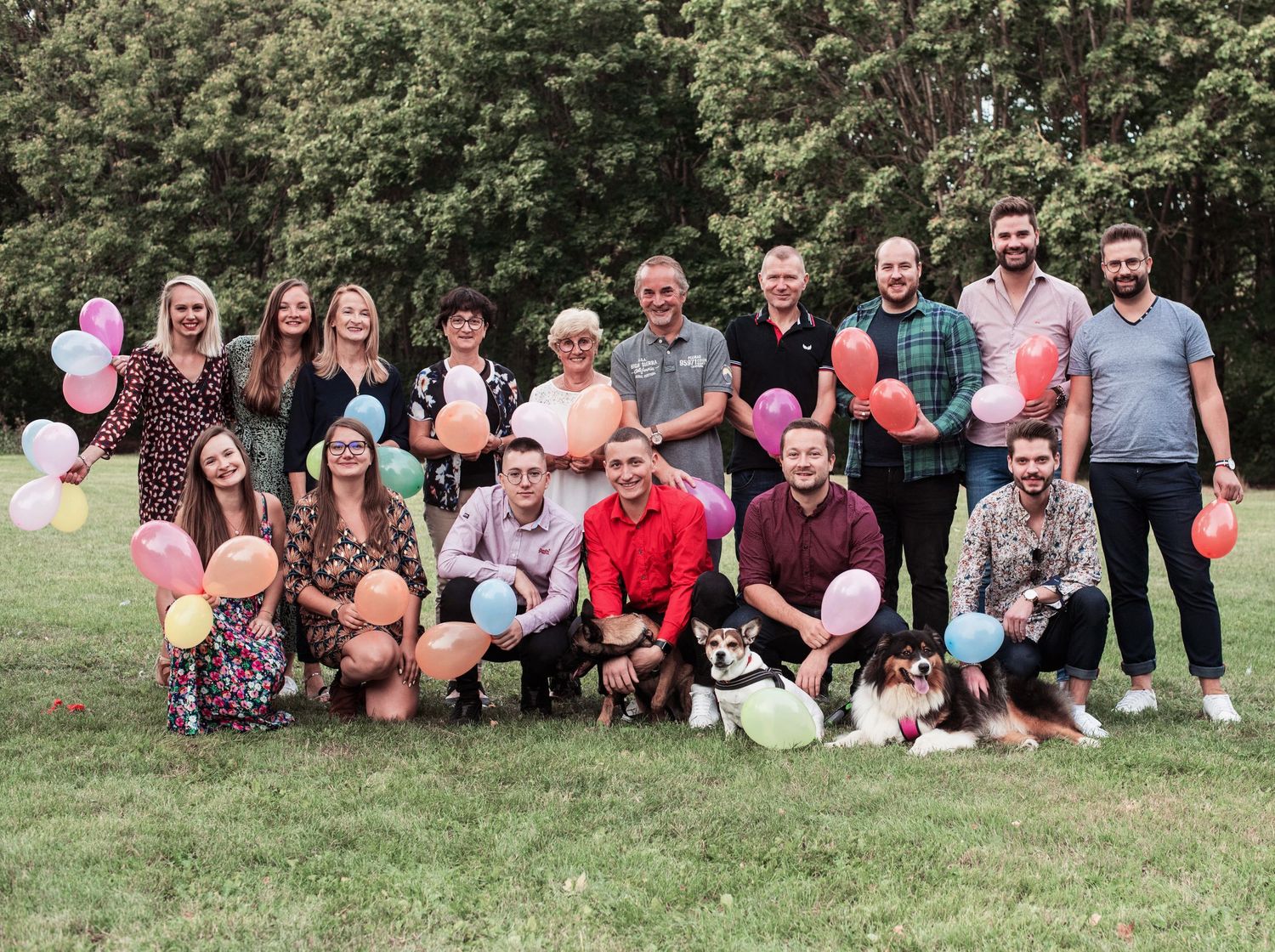 Large group photo with people sitting on grass holding colorful balloons in a park setting.