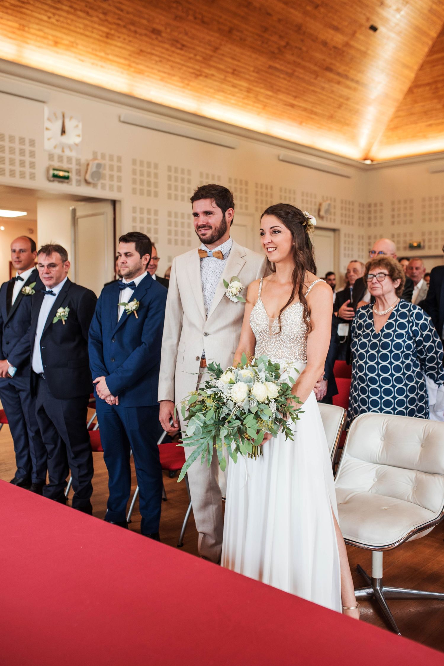 Wedding party lines up in formal attire against a wood-paneled wall with red carpet.