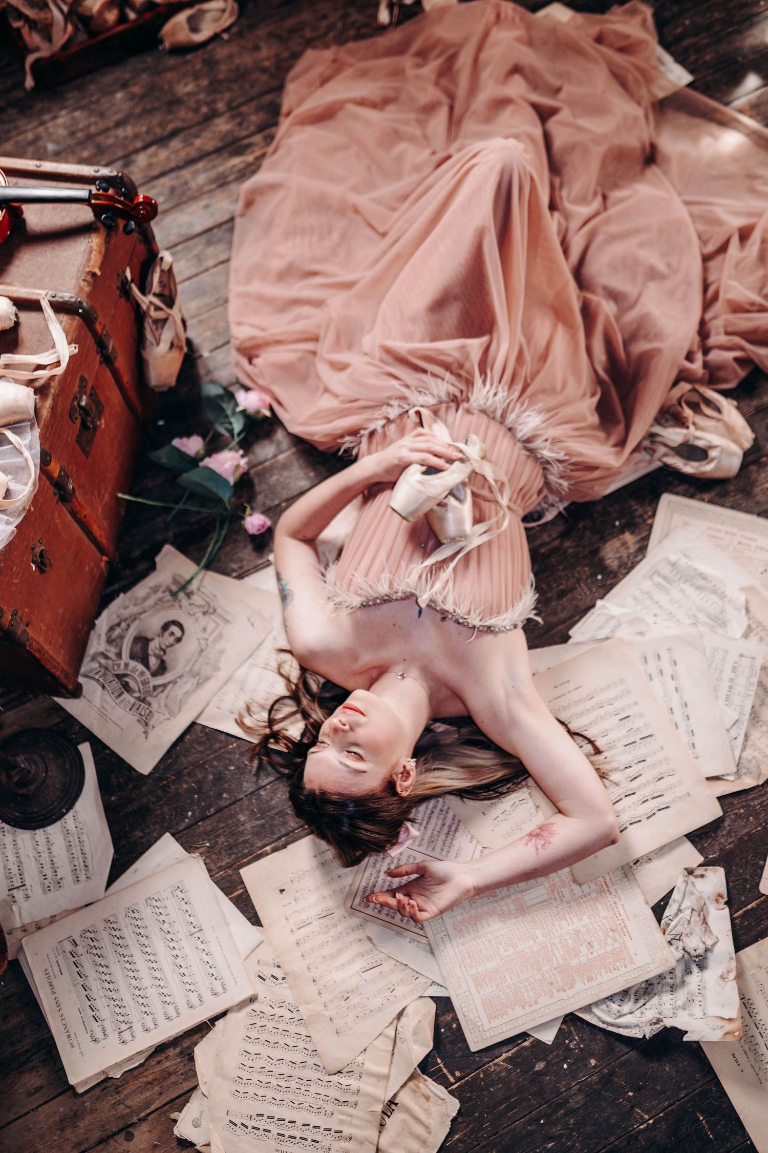 Ballerina in pink dress lying on wooden floor surrounded by scattered sheet music and ballet pointe shoes.