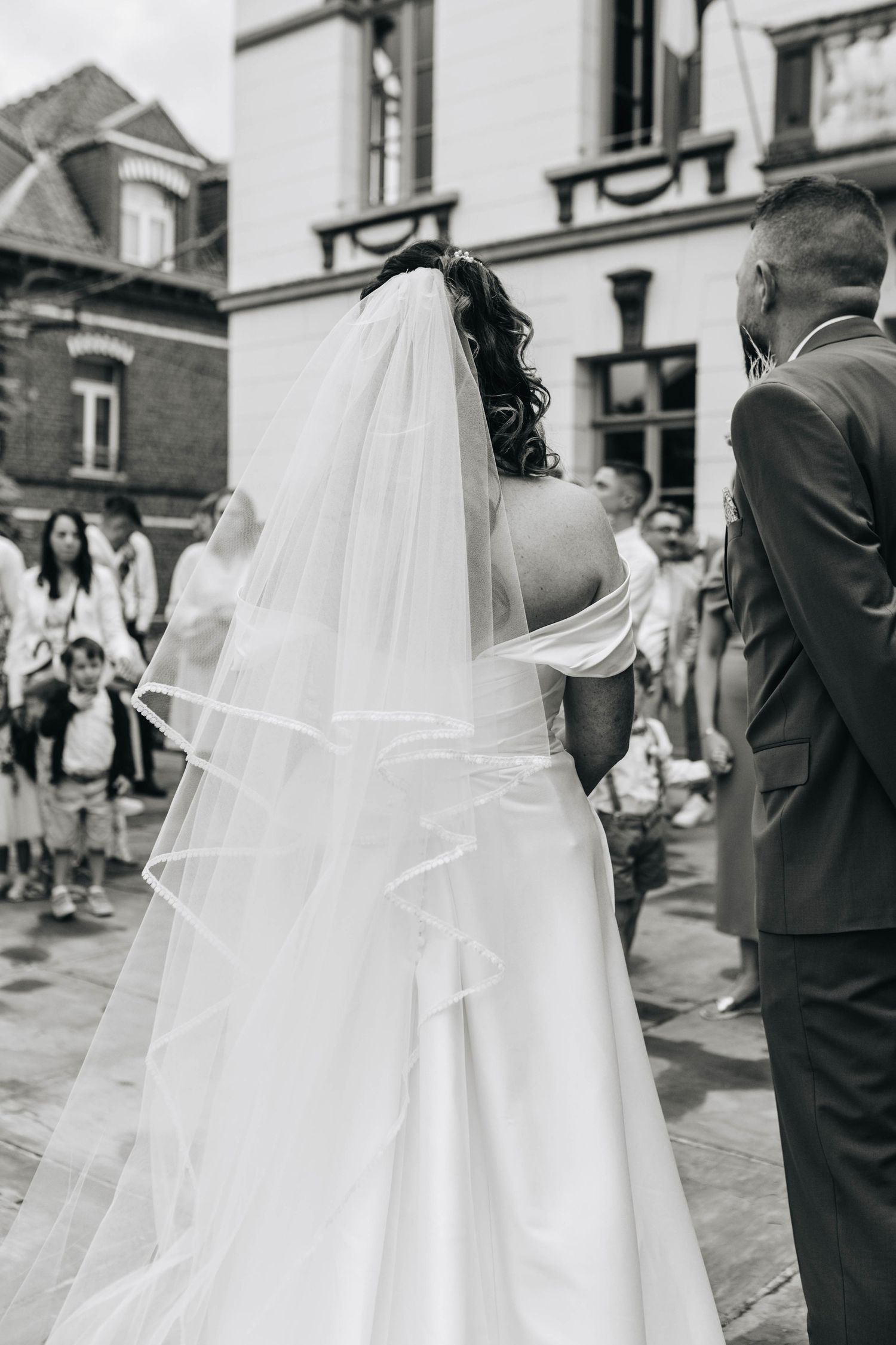 A flowing white wedding veil trails behind as the couple walks down a city street.