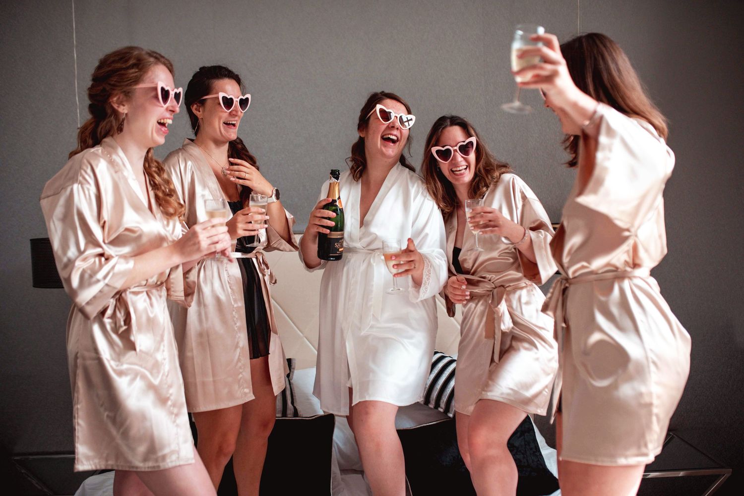 Bridesmaids in matching silk robes celebrating with drinks and laughter during pre-wedding preparations.