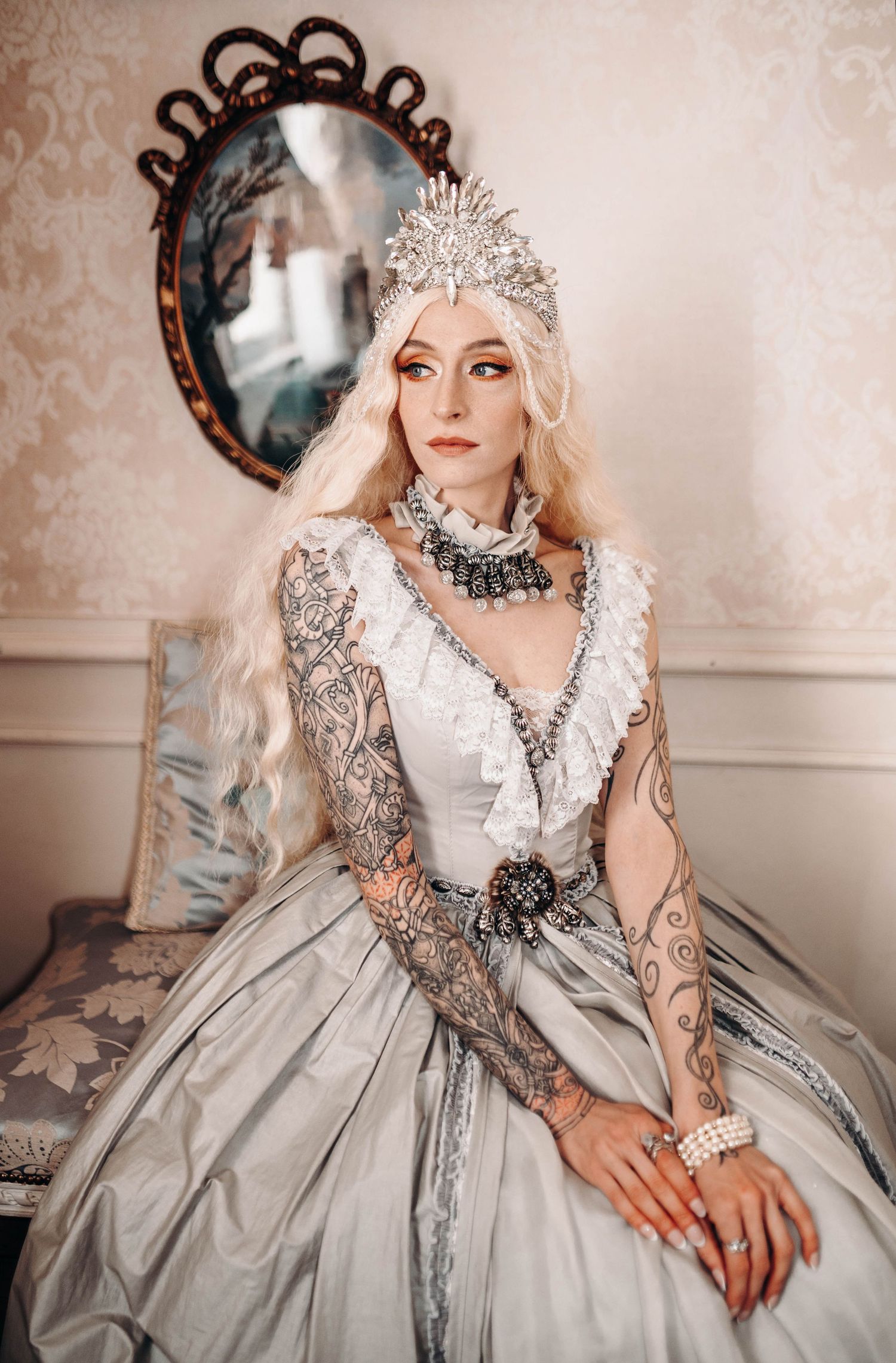A tattooed woman in a silver ball gown and crystal crown with long white hair poses regally before an ornate mirror.