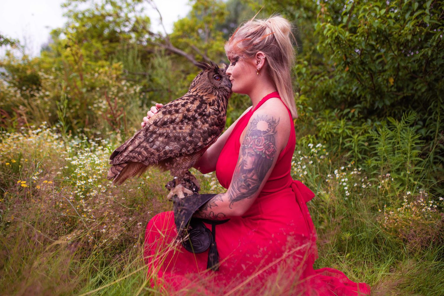 An eagle owl spreads its wings while perched on someone wearing a flowing red dress in a meadow.