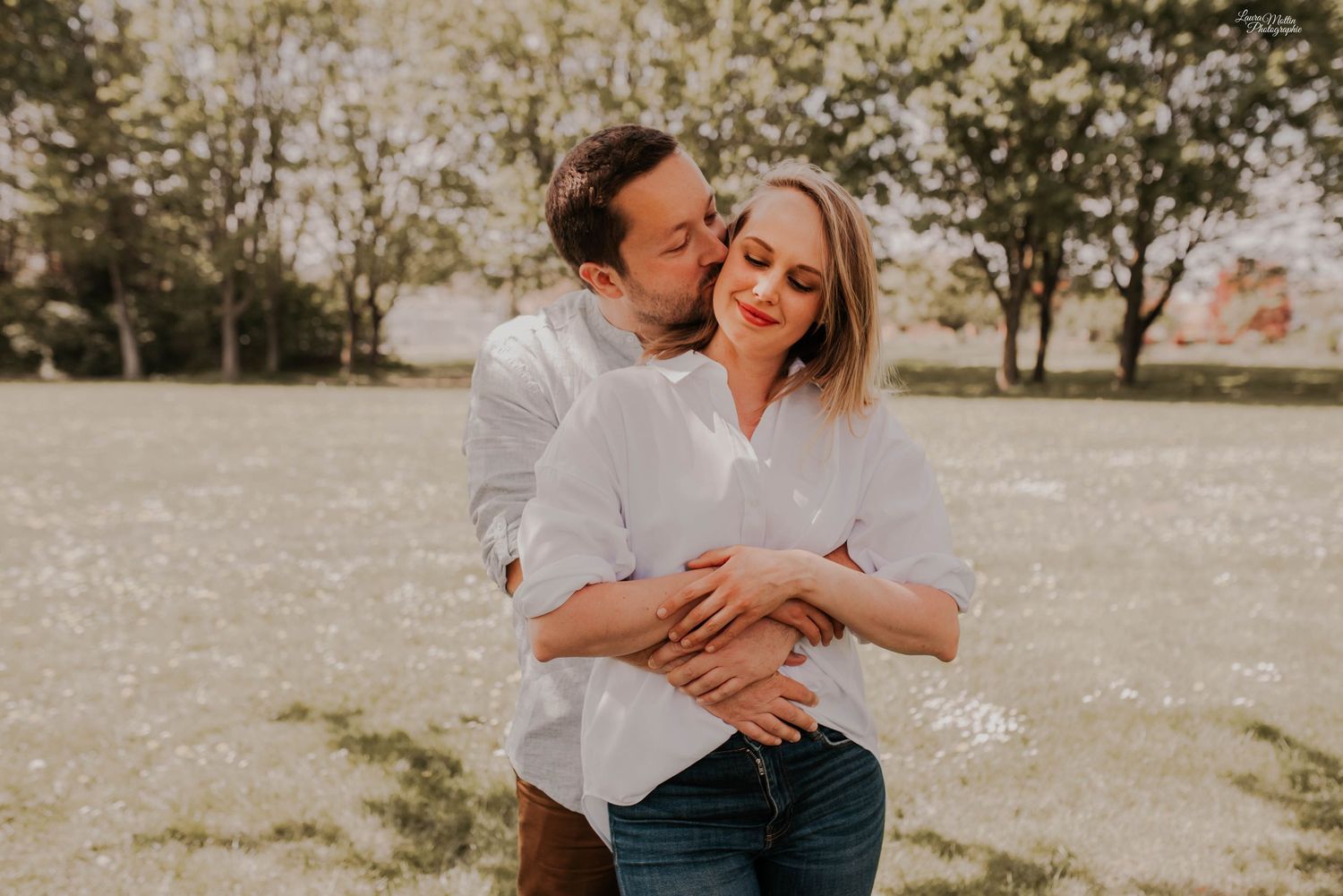 Romantic couple embraces in a sunlit field with trees in the background during an engagement photoshoot.