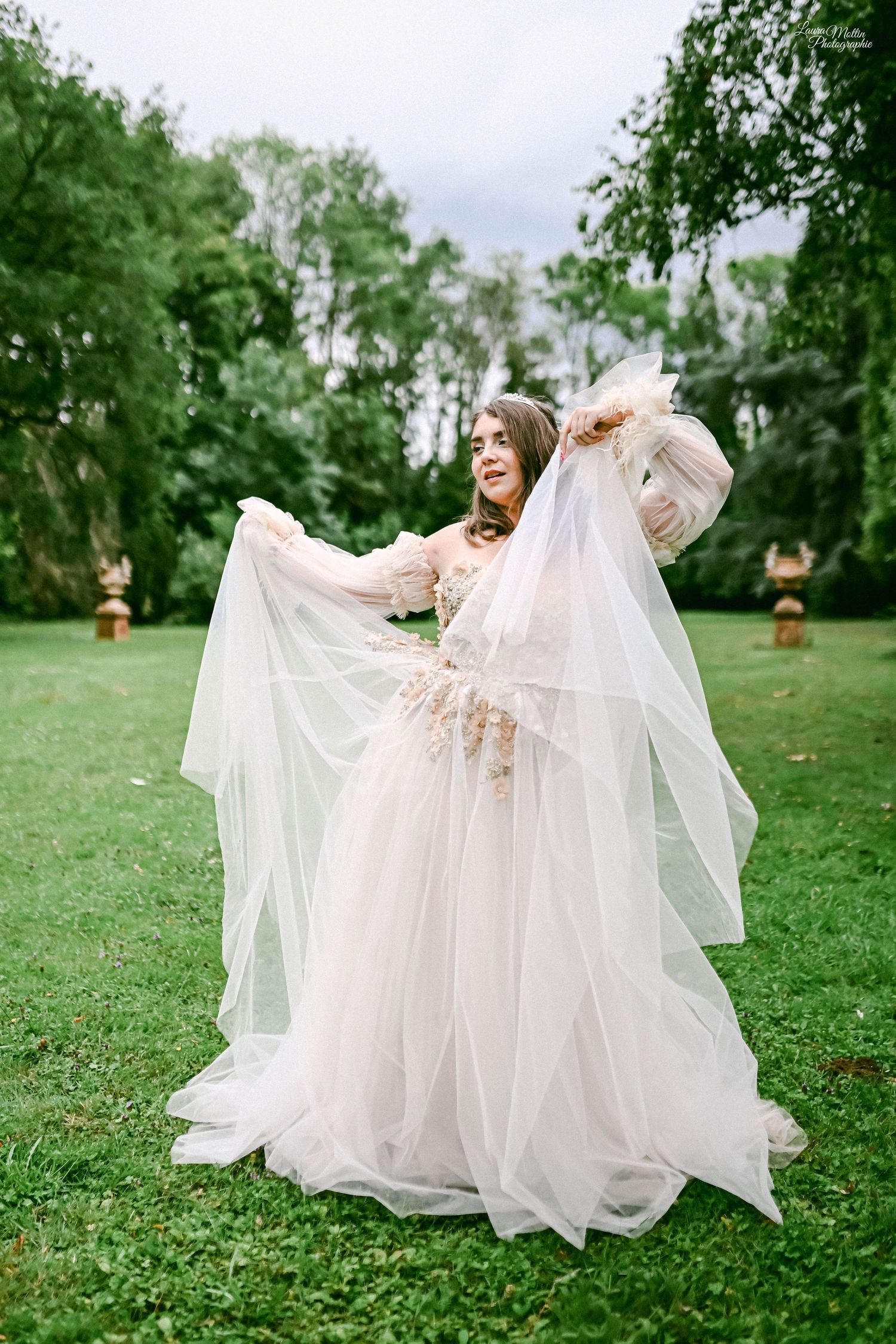 Bride in delicate tulle gown poses dramatically in garden with tall trees.