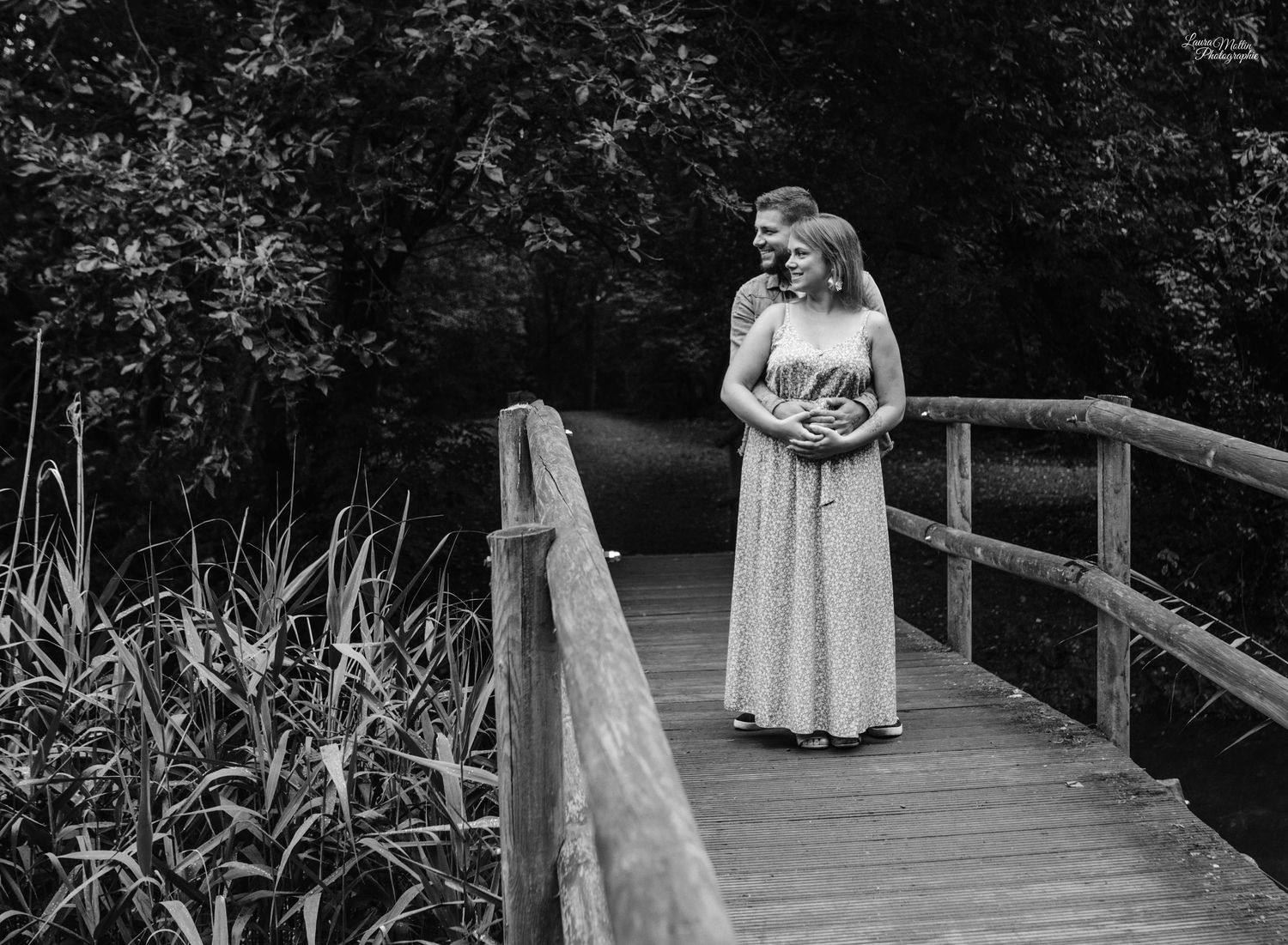 Black and white photo of couple embracing on wooden bridge surrounded by nature.