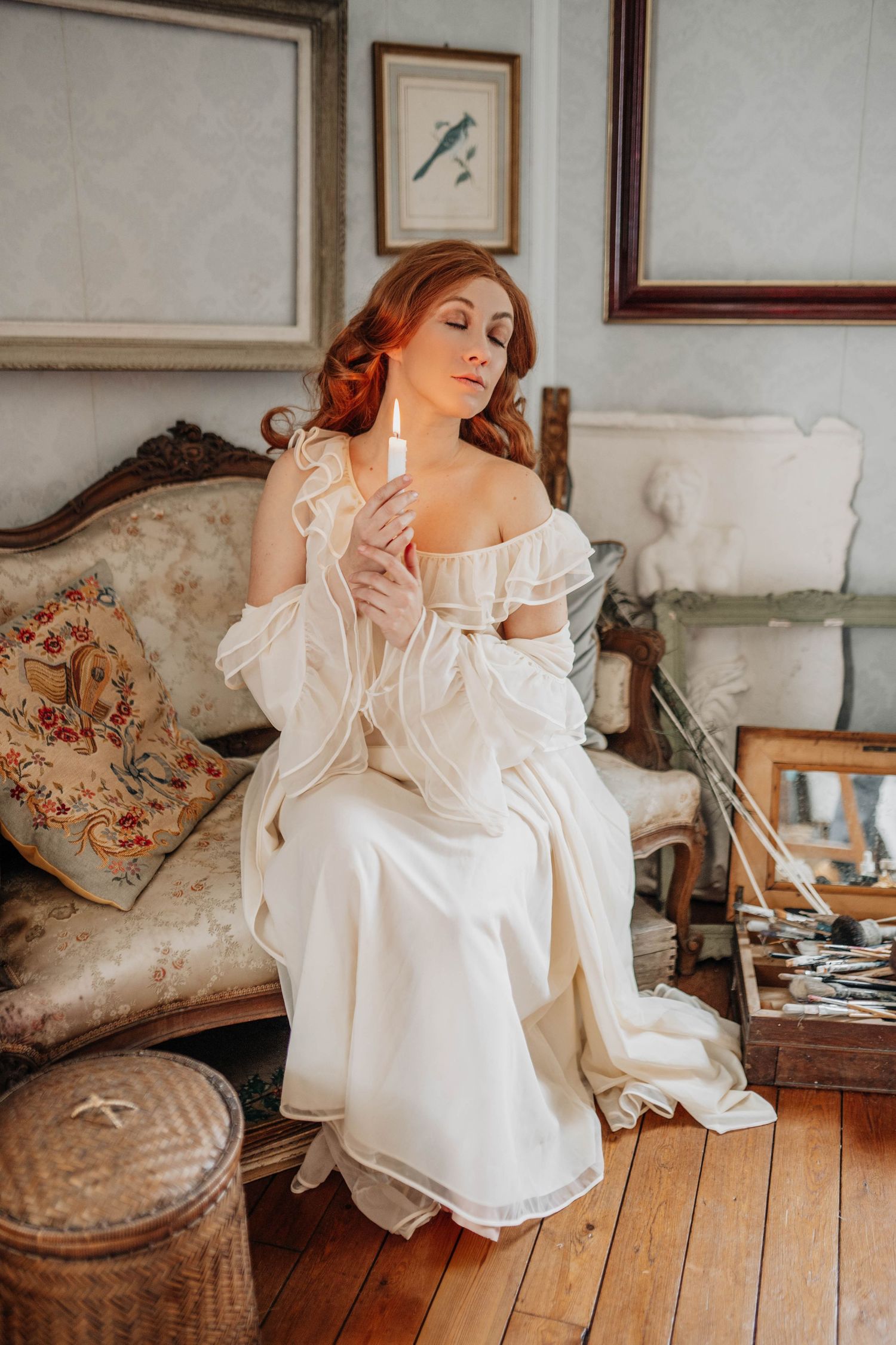 A red-haired woman in a white vintage gown holds a candle while seated on an ornate antique sofa.