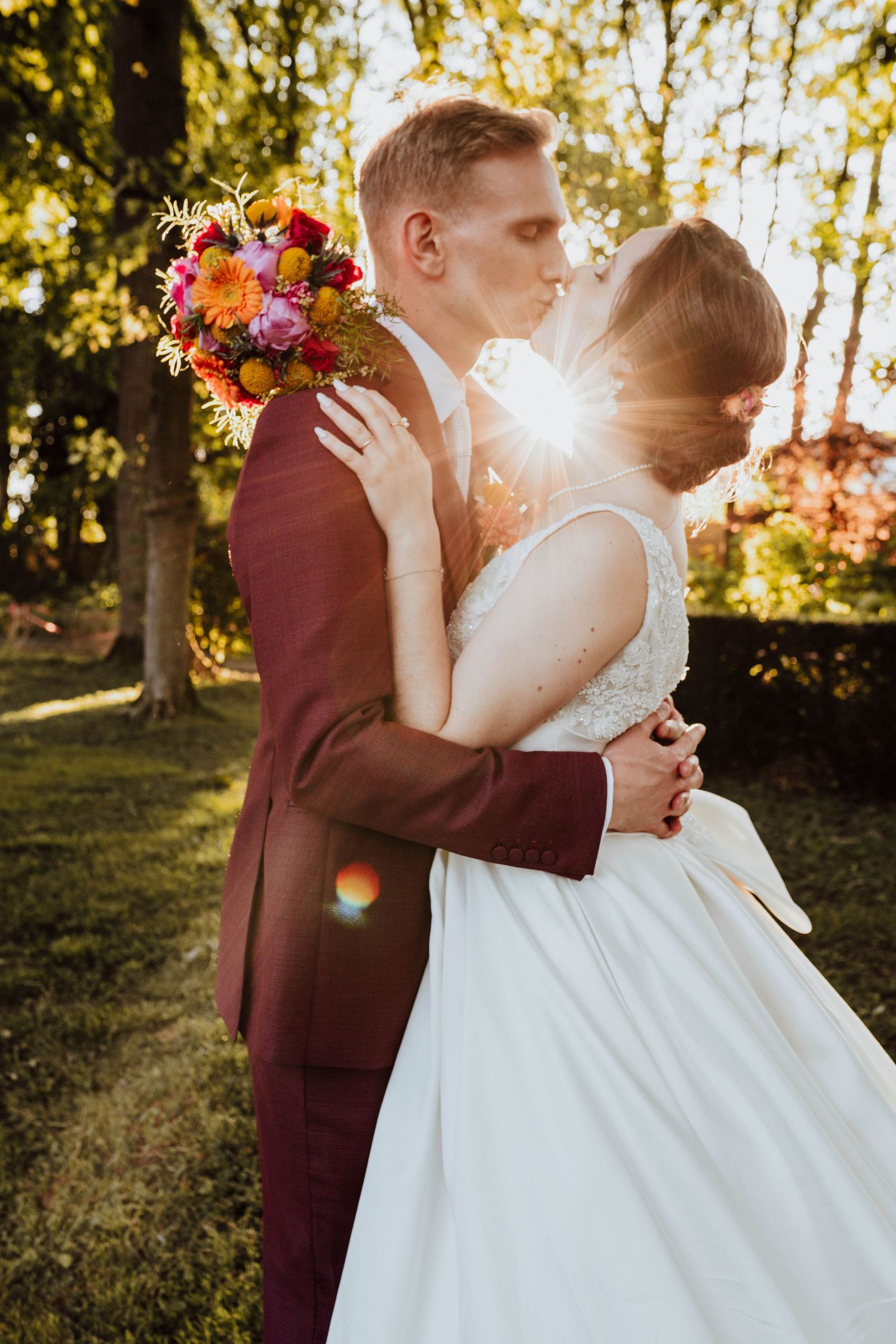 Bride and groom share a kiss outdoors in golden sunlight, she in white gown holding colorful bouquet, he in burgundy suit.