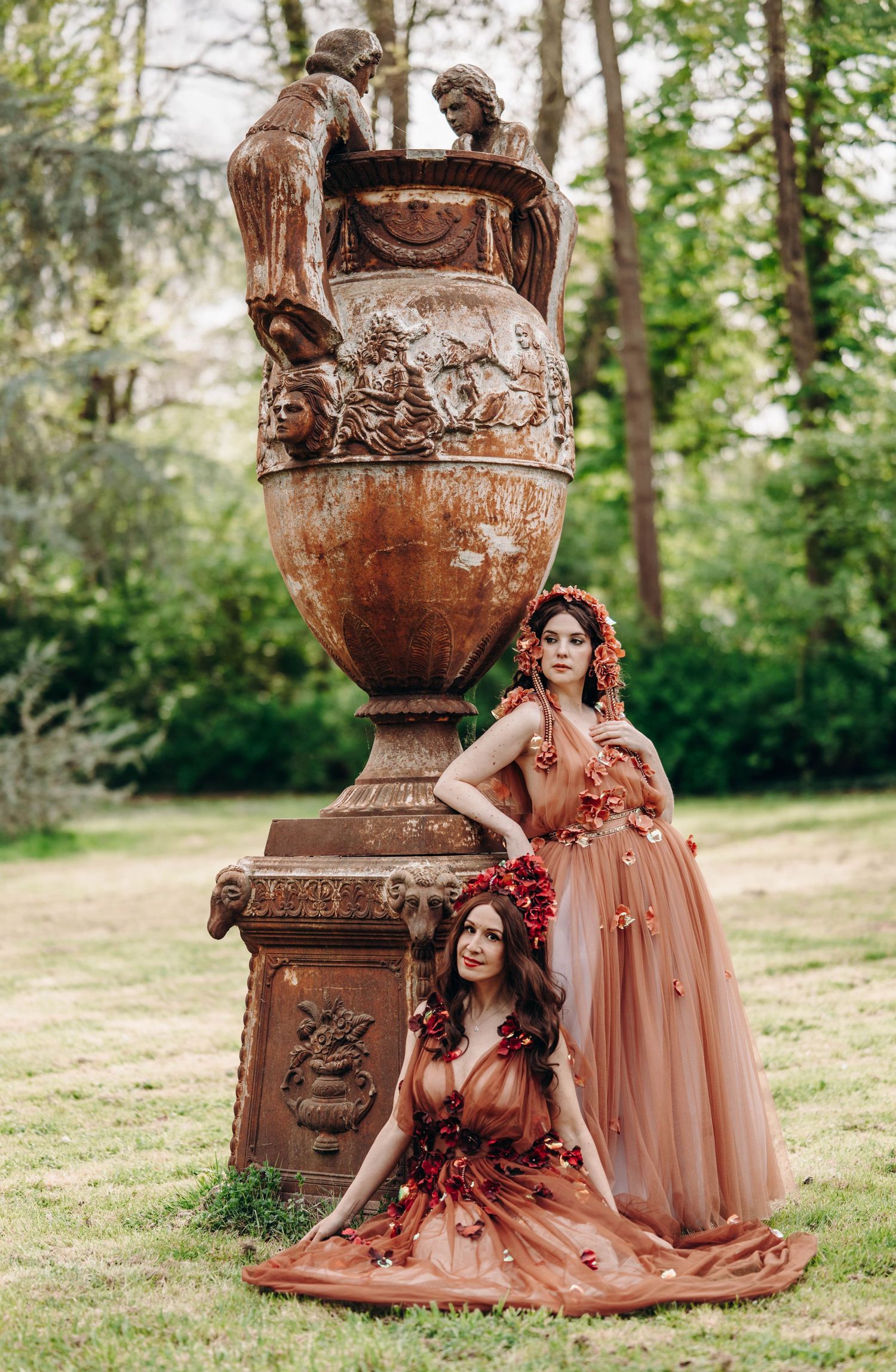 Two women in rust-colored floral gowns pose elegantly beside a large ornate stone urn in a lush garden.