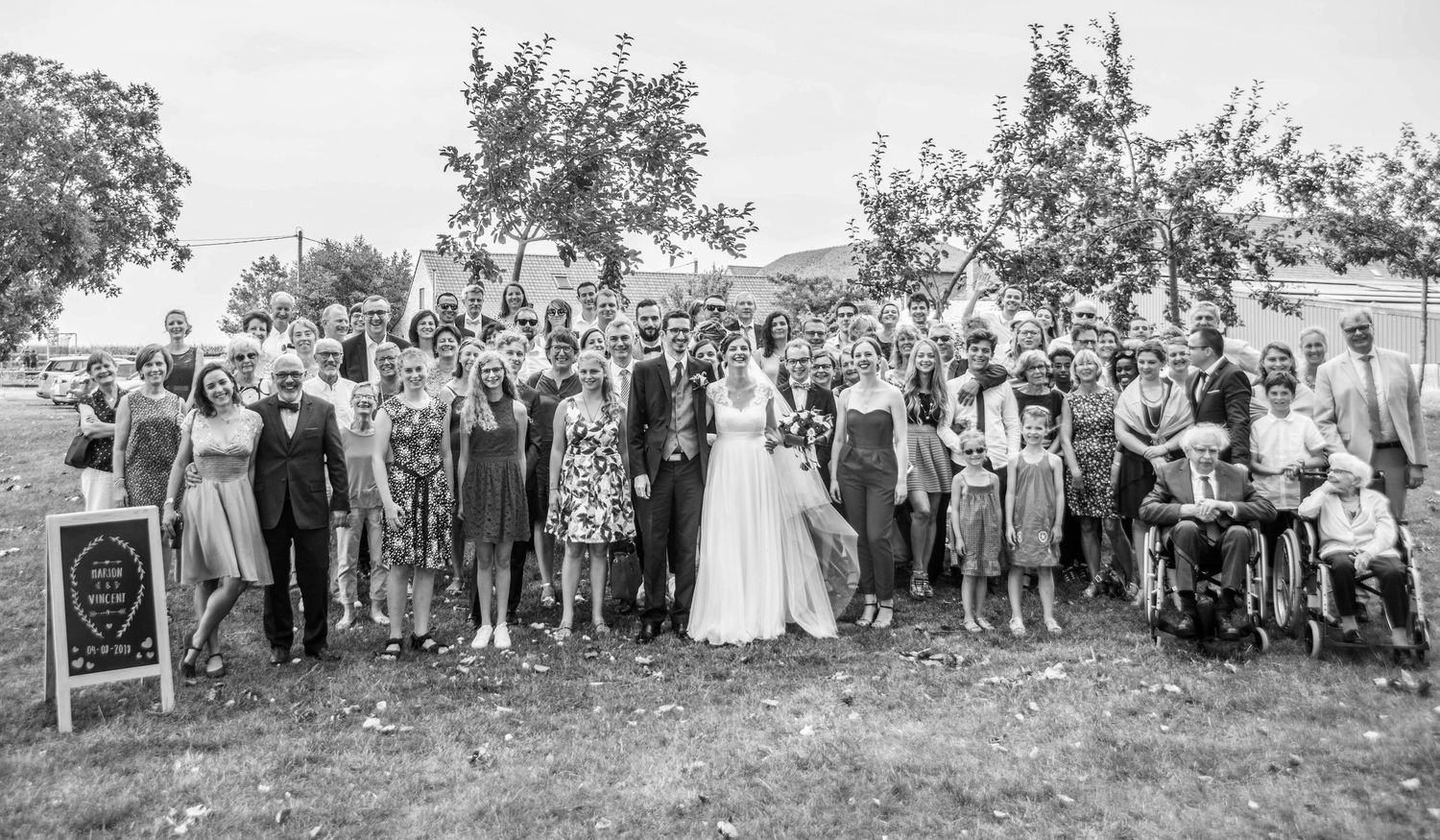 Large group wedding photo of guests and newlyweds gathered outdoors under trees.