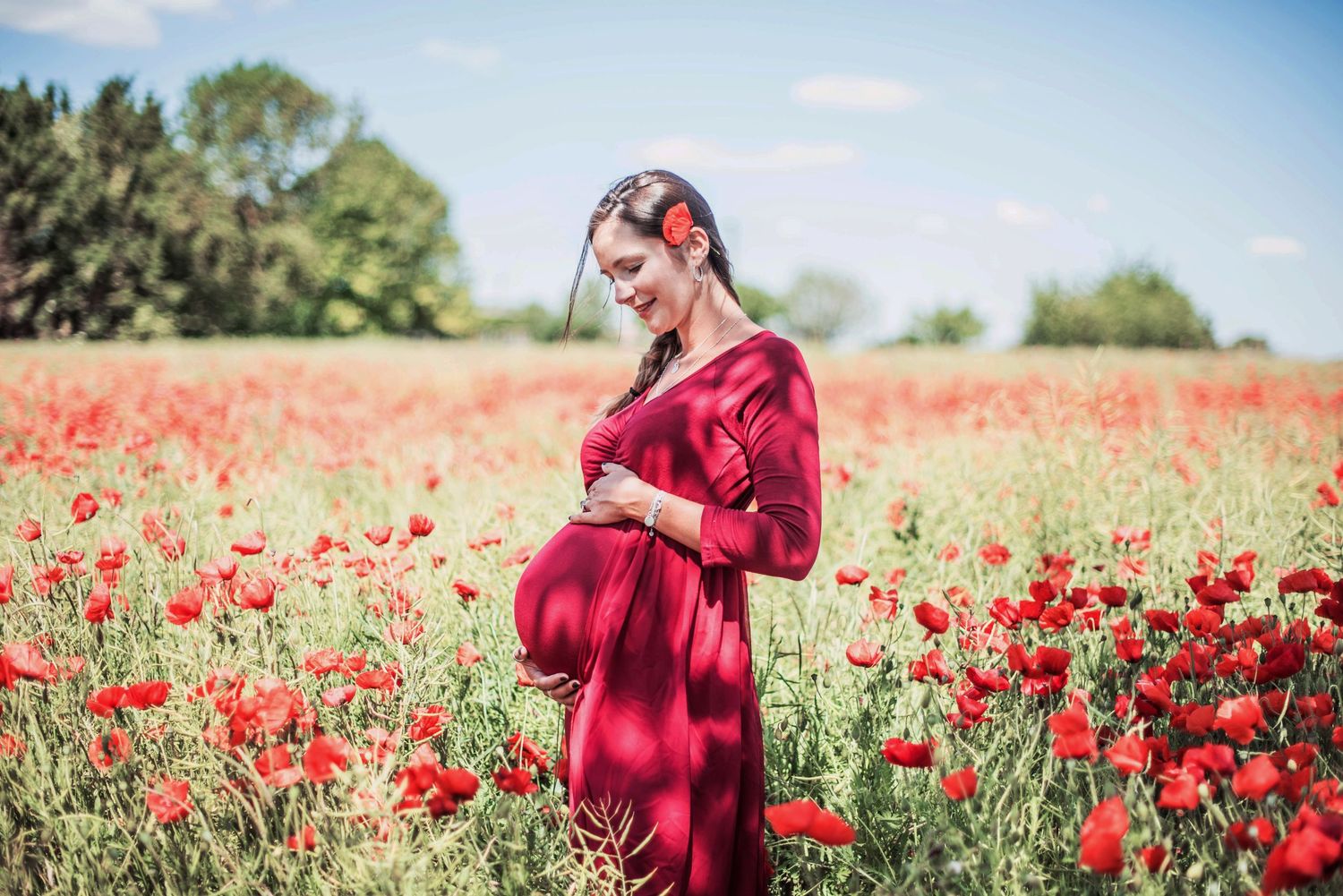 Maternity photography in red dress standing among vibrant poppy field under blue sky.