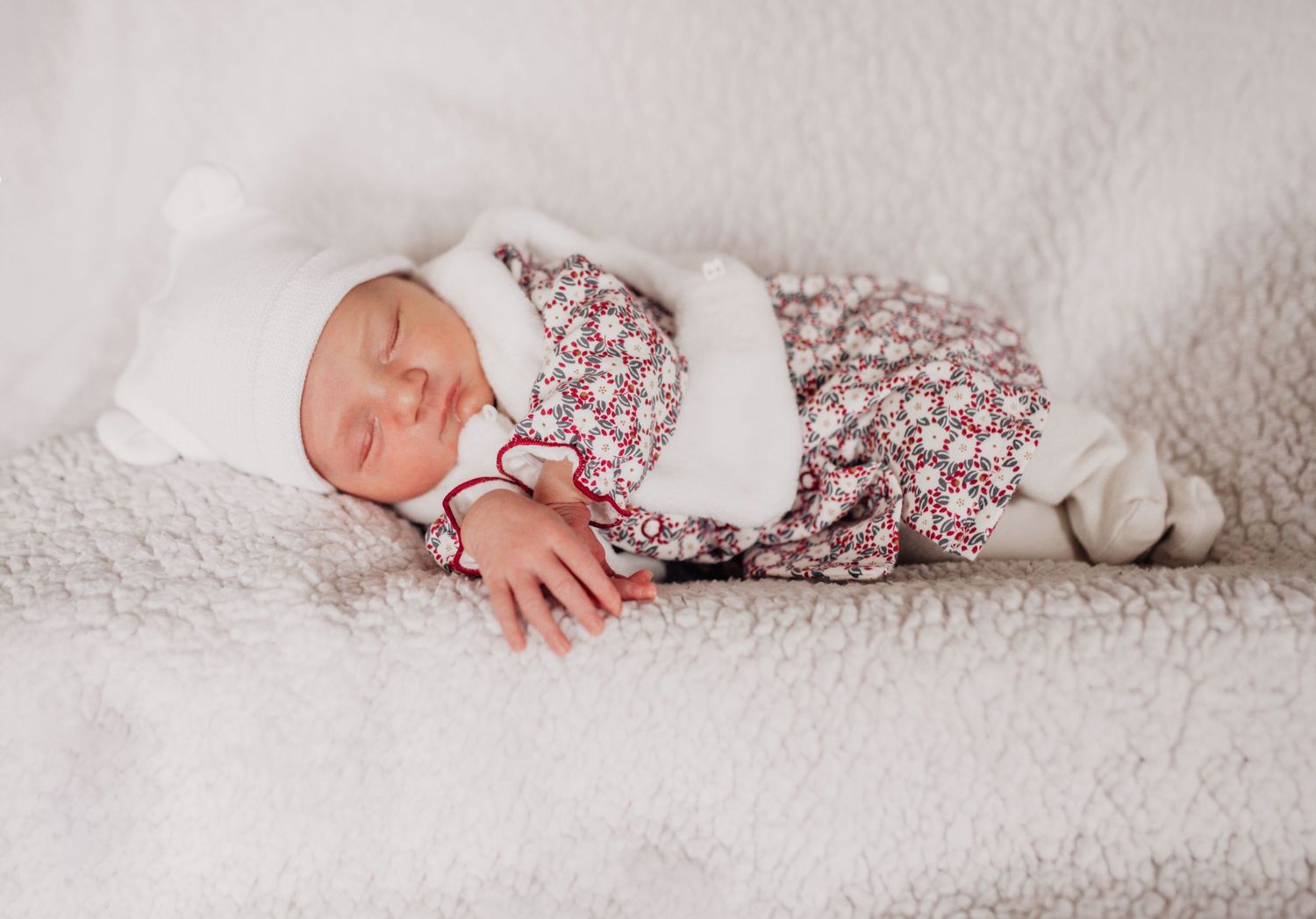 Newborn baby sleeping peacefully on white textured blanket wearing floral outfit.