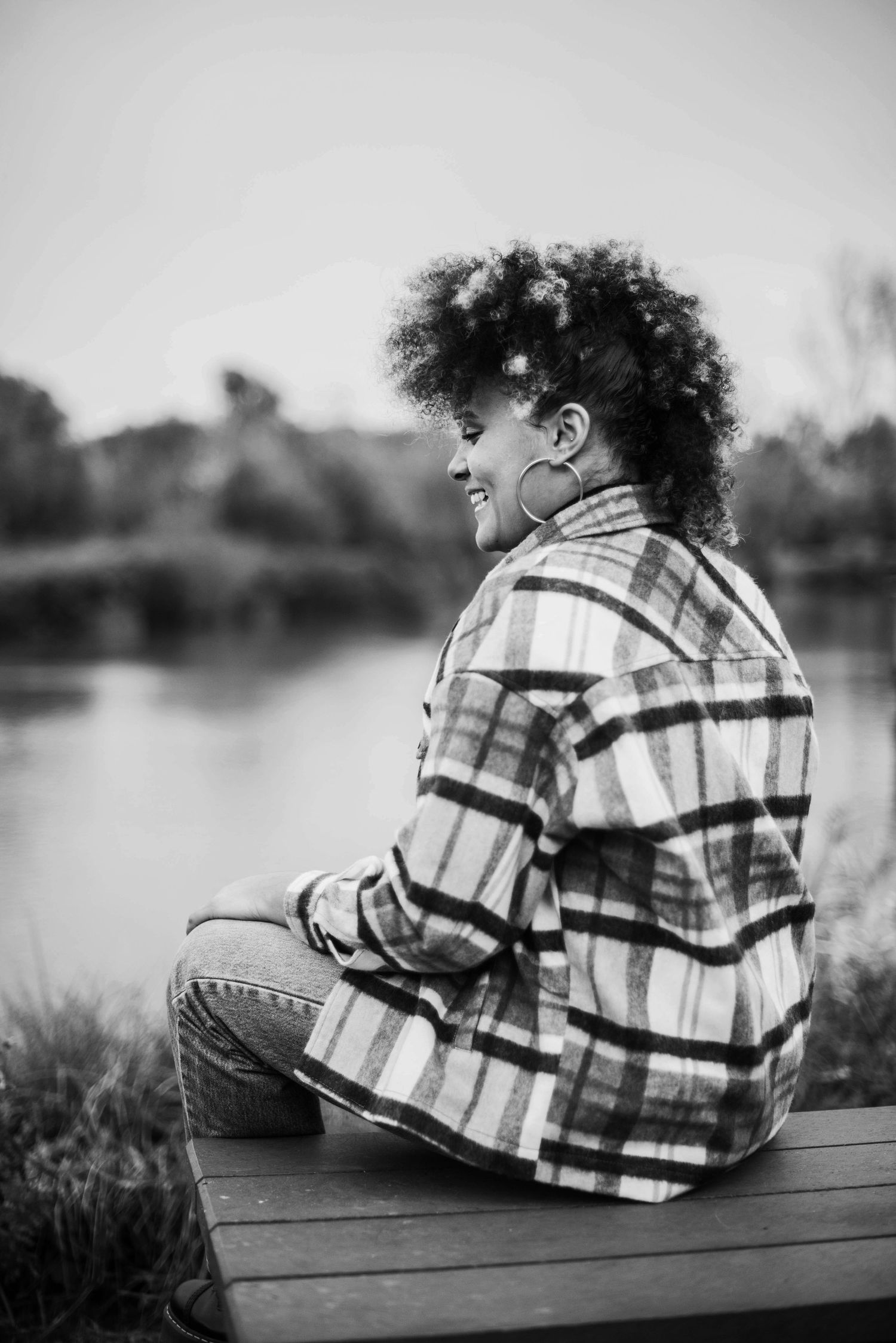 Person in plaid shirt relaxing on wooden bench by a lake in black and white.