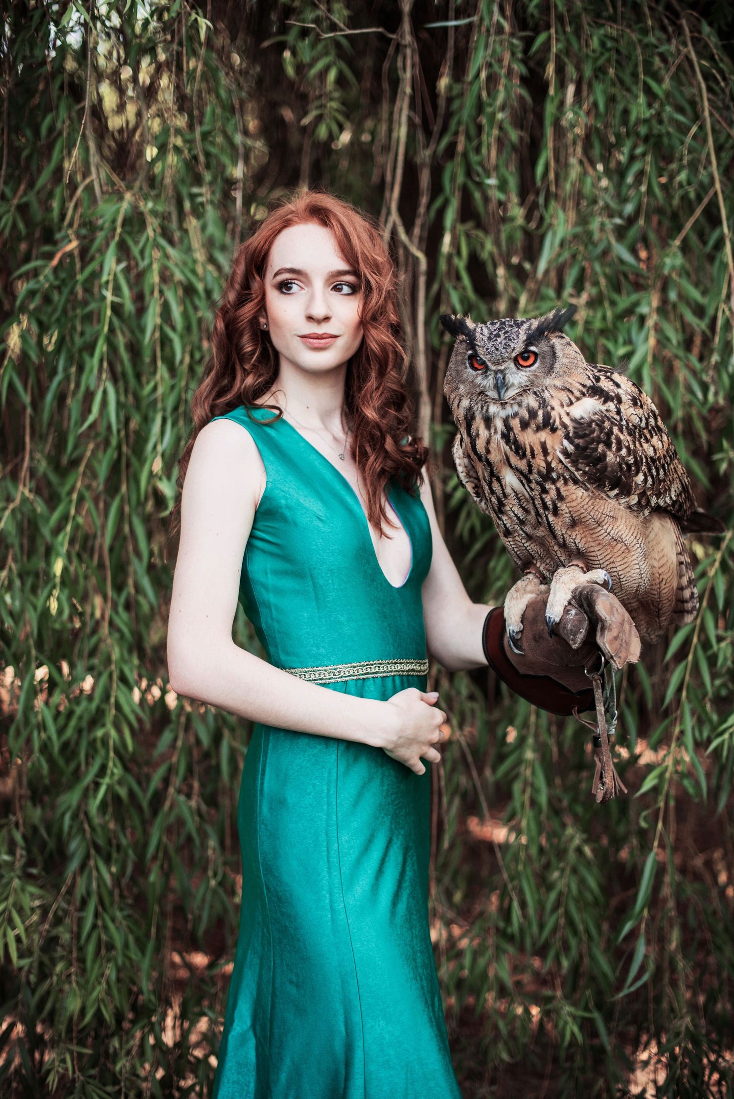 Person in emerald green dress holding a large owl against willow tree backdrop.