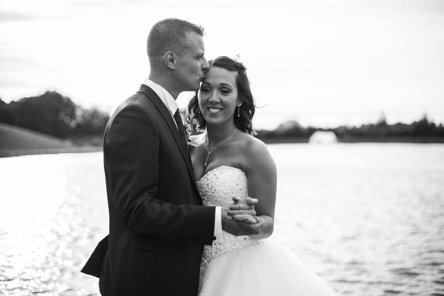 A romantic black and white wedding photo of a couple embracing by a lake.