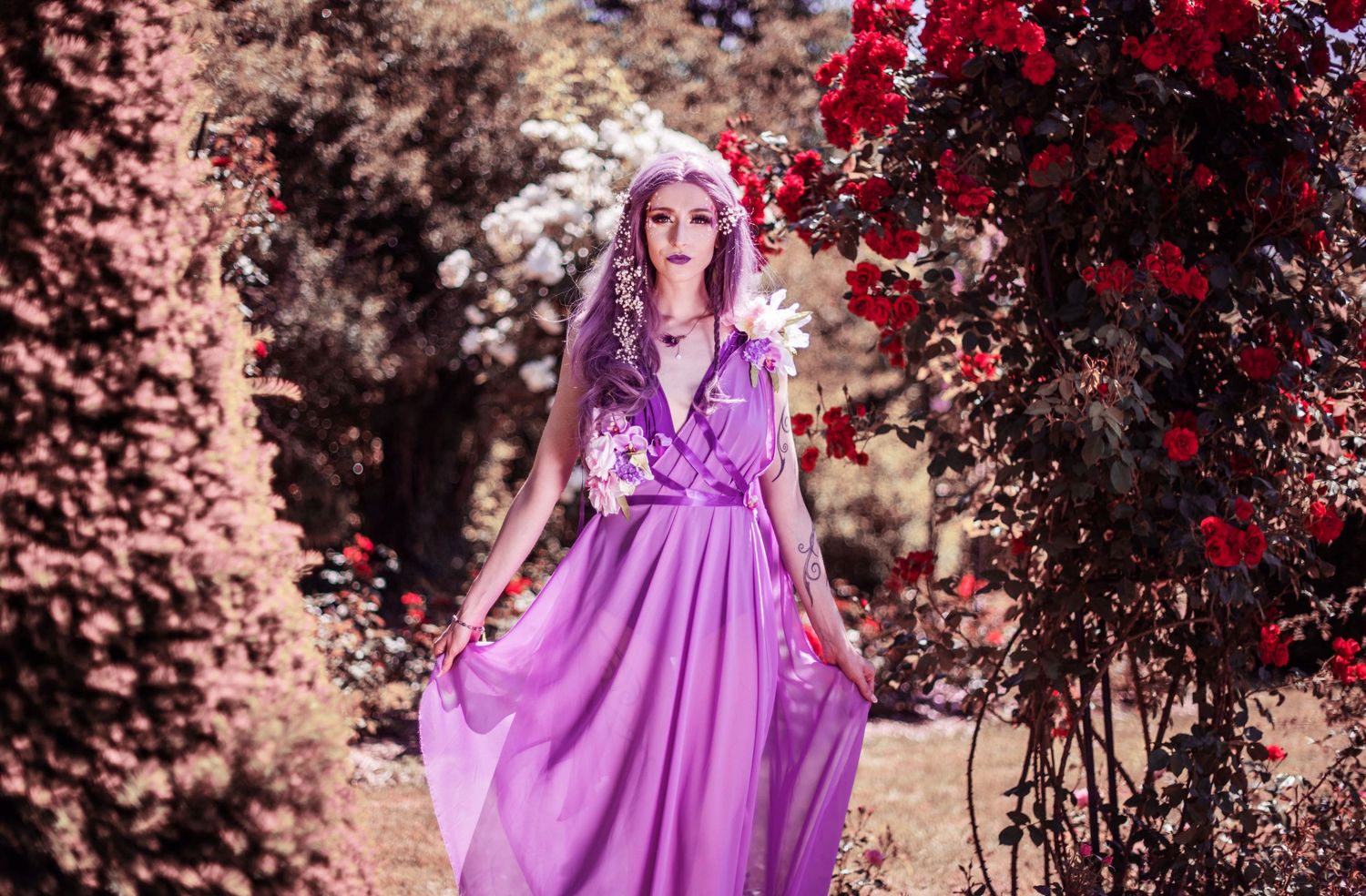 A person in a flowing purple gown poses among red flowers and foliage in a dreamy outdoor setting.
