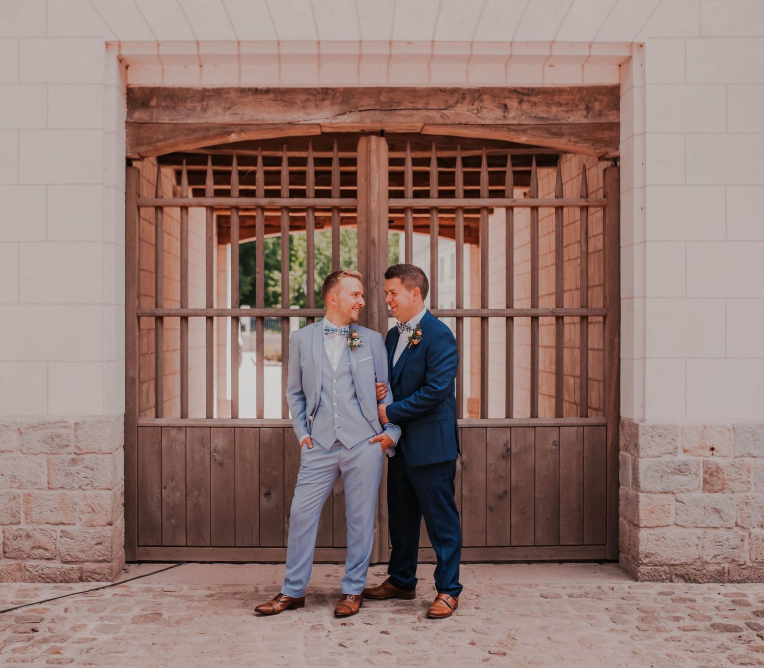 A couple in contrasting suits poses together in front of a rustic wooden gate.