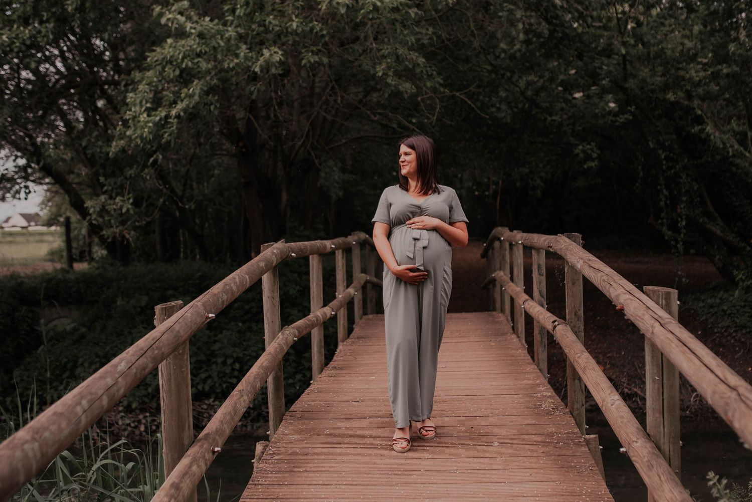 Pregnant woman in flowing dress stands alone on wooden bridge surrounded by trees.