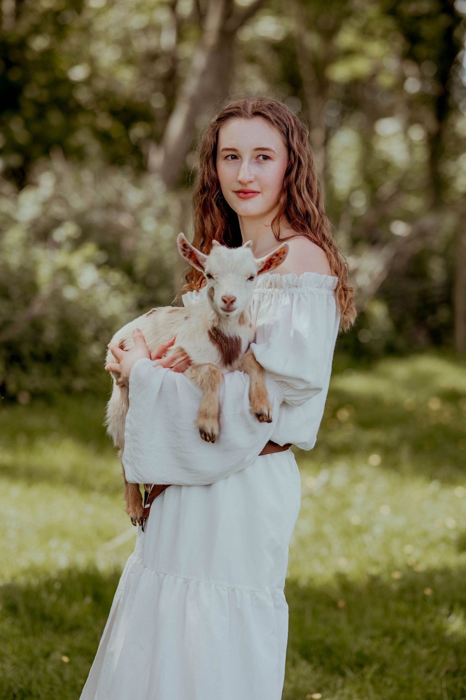 Young woman in white off-shoulder dress holding a baby goat in a lush green outdoor setting.