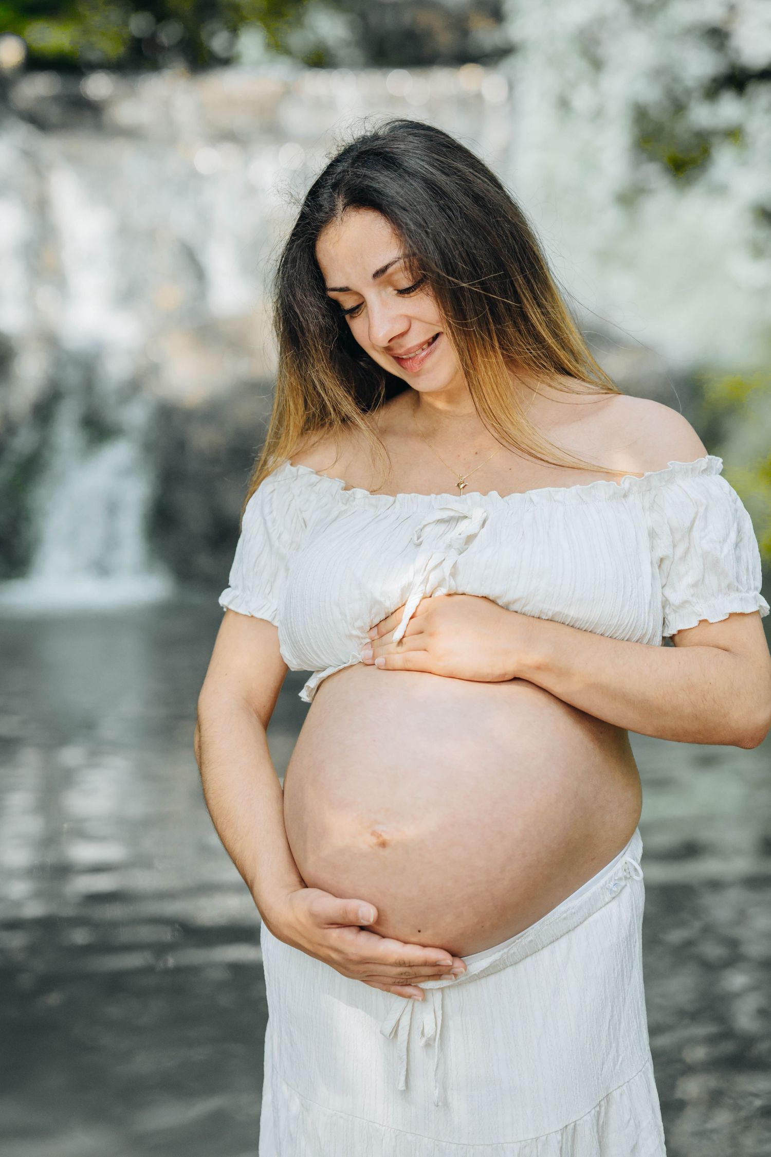 Smiling pregnant woman in white off-shoulder dress cradling her bare belly near a serene waterfall backdrop.