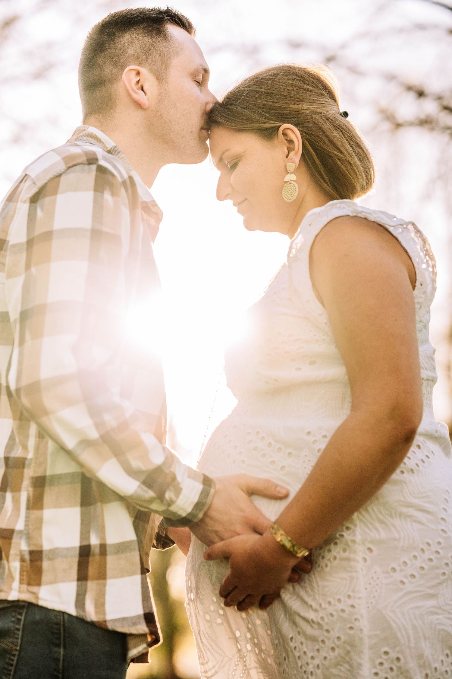 Couple sharing a tender moment outdoors, man kissing pregnant woman's forehead in golden sunlight.