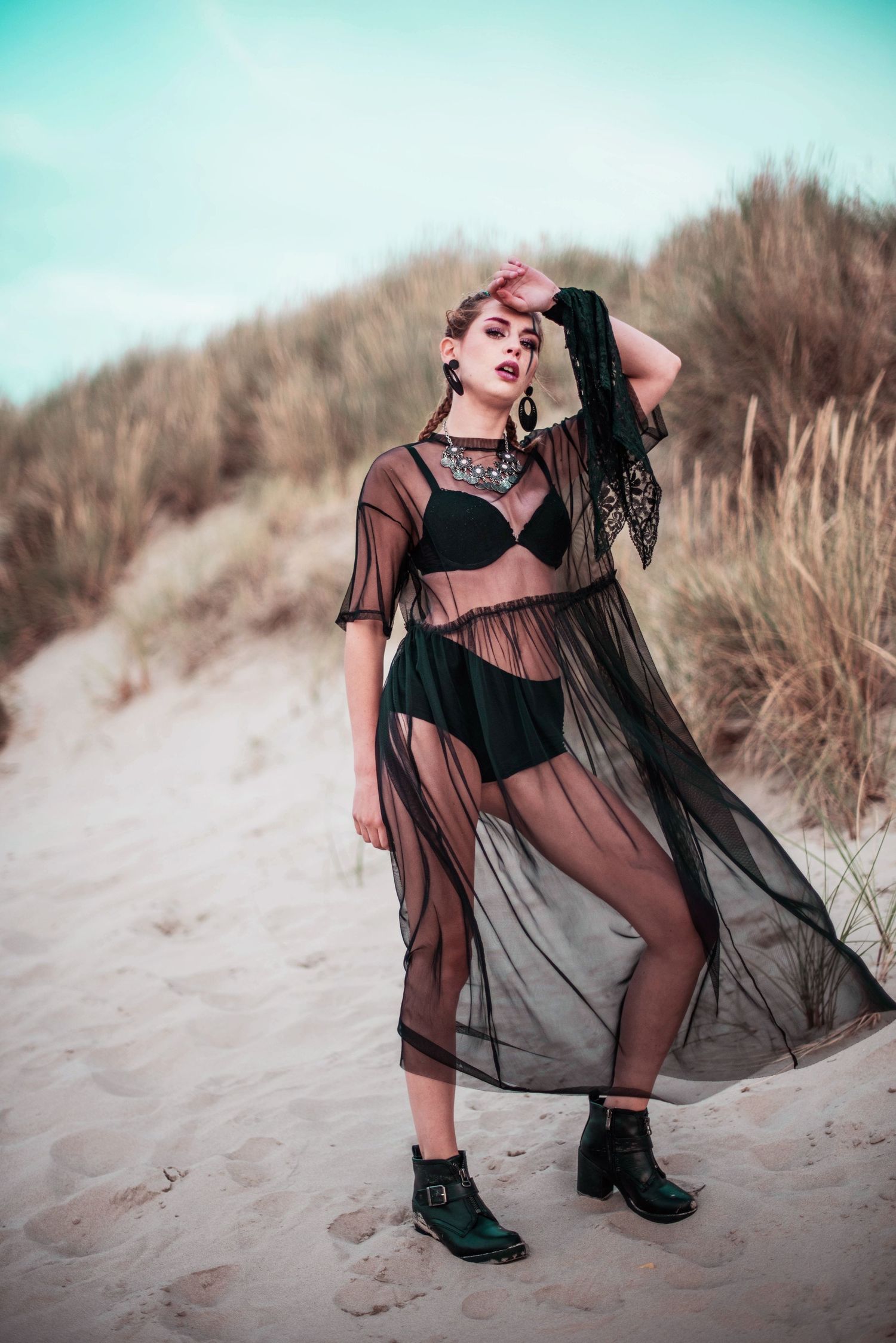 Model in black sheer dress and boots poses on beach dunes with turquoise sky.